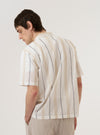 A model wears the Ecru/Navy/Sand Hendrix Stripe Road Shirt over a light-colored top, shown from a rear three-quarter angle against a plain white background. The shirt features a vertical multi-colored stripe pattern in ecru, navy, and sand, with a straight hem and short sleeves. The camp collar is visible from the side.