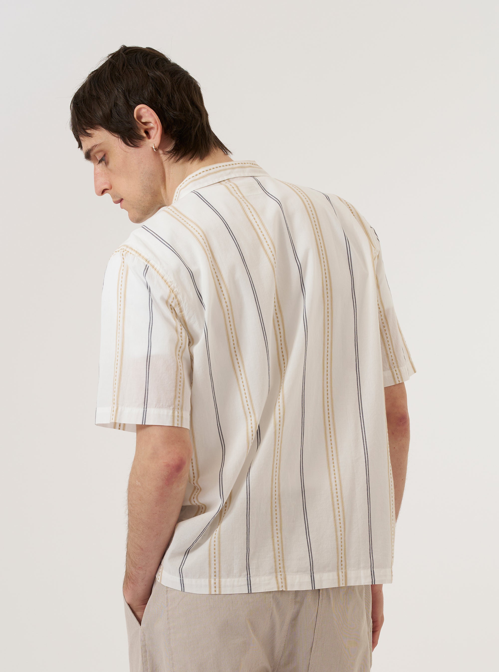 A model wears the Ecru/Navy/Sand Hendrix Stripe Road Shirt over a light-colored top, shown from a rear three-quarter angle against a plain white background. The shirt features a vertical multi-colored stripe pattern in ecru, navy, and sand, with a straight hem and short sleeves. The camp collar is visible from the side.