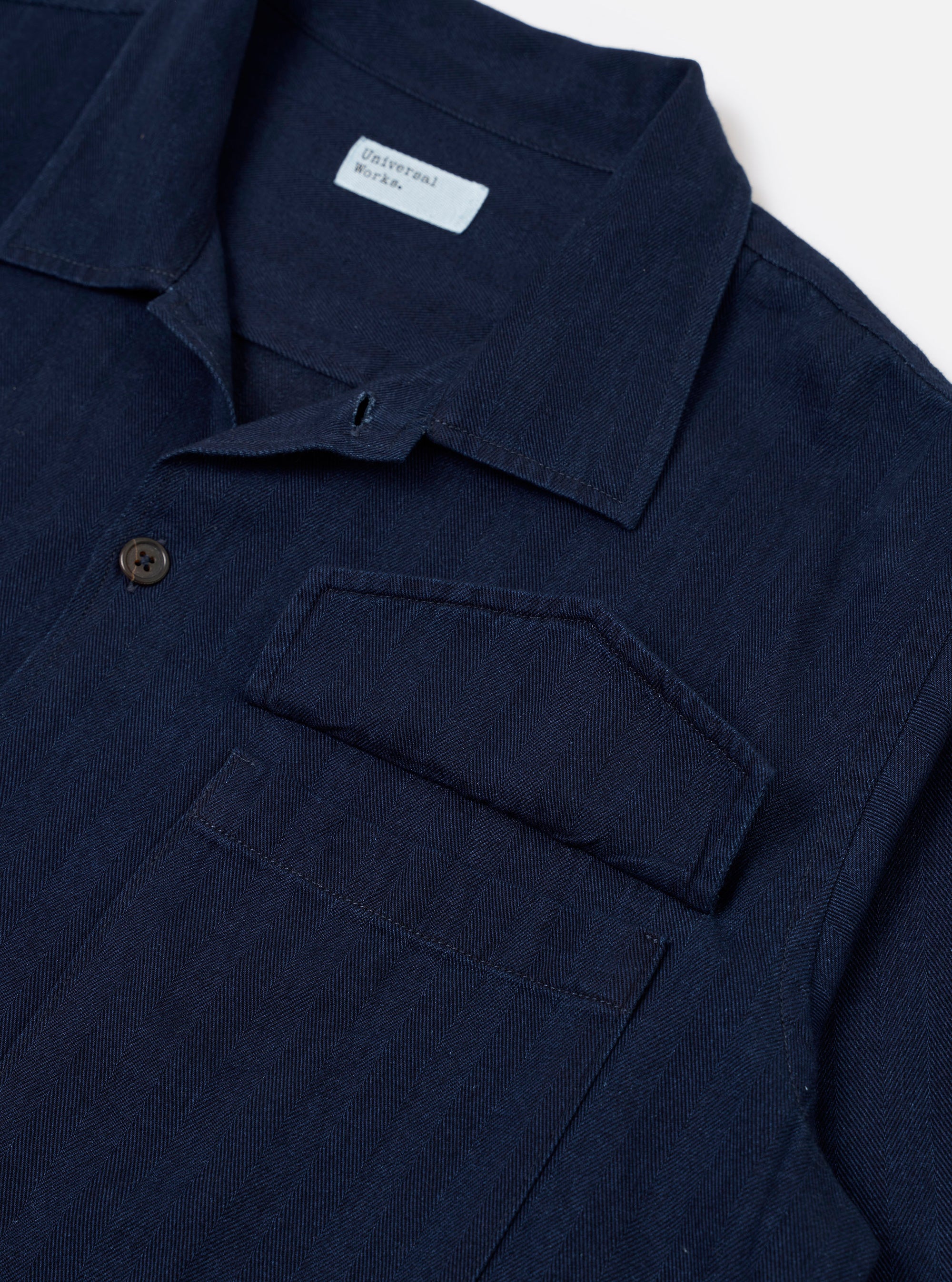 The Indigo Recycled Herringbone Attendant Shirt is worn by a model against a plain, light-colored background. The shirt is shown from a three-quarter rear angle, highlighting the straight hem, the back yoke, and the utility-style construction. The indigo-dyed fabric features a visible herringbone weave. The model is also wearing olive-colored trousers.