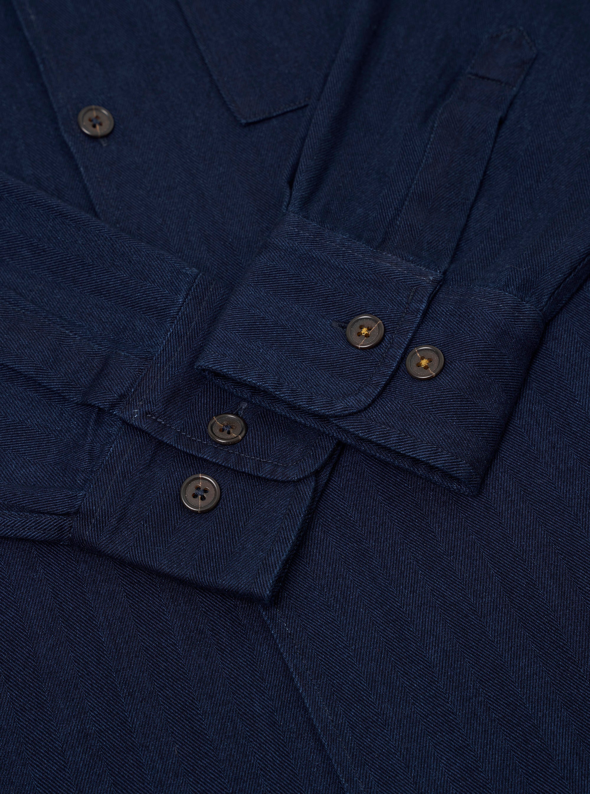 The Indigo Recycled Herringbone Attendant Shirt is shown from the back as a flat lay against a white background. The shirt features a straight hem, a single horizontal yoke across the shoulders, and a simple flat collar. The indigo fabric has a visible vertical herringbone texture throughout.