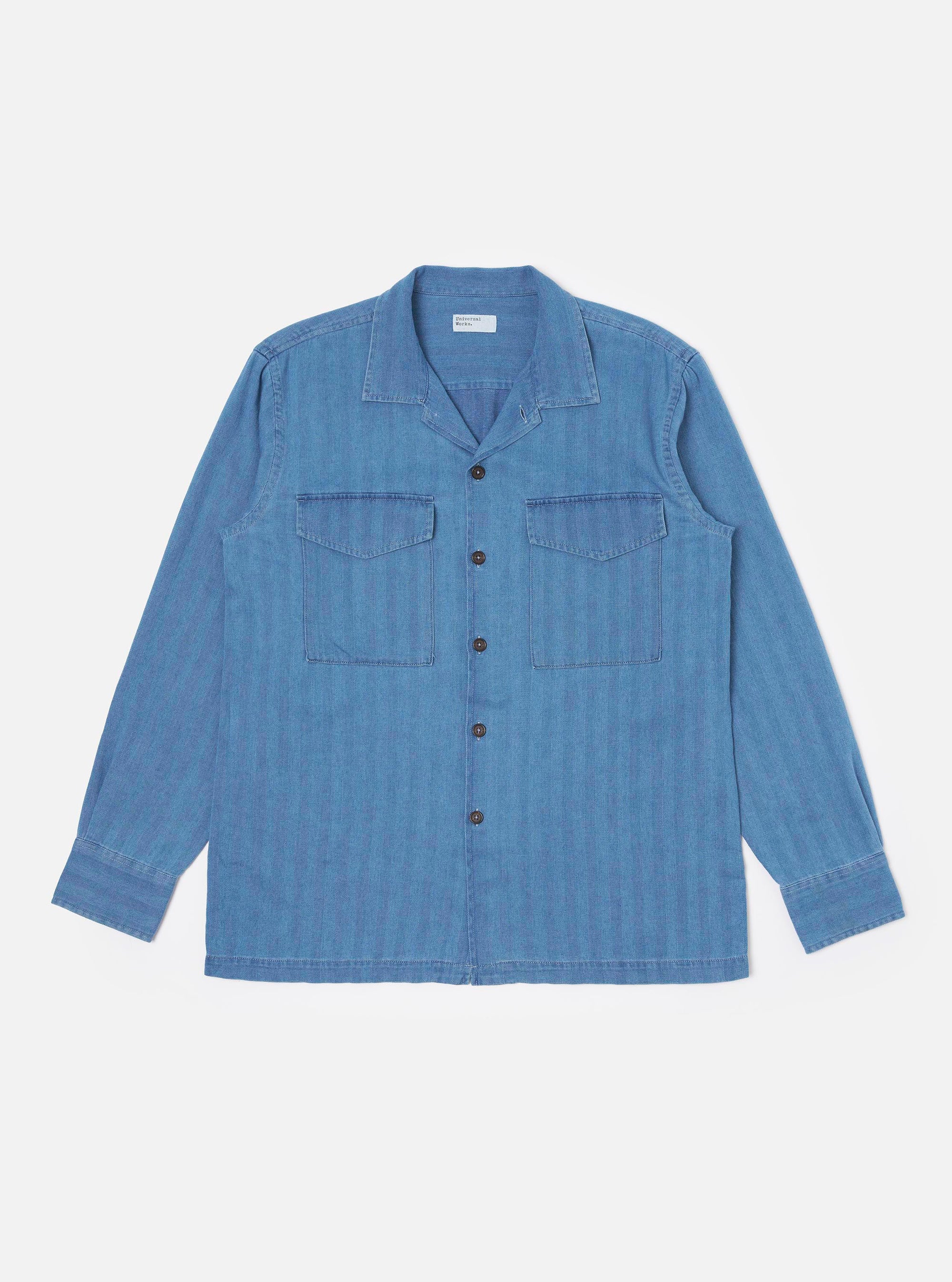 The Washed Indigo Recycled Herringbone Attendant Shirt is shown as a flat lay against a plain white background. The shirt features a flat collar, a full-length button-down front, and two large chest pockets with button-closure flaps. The washed indigo blue fabric displays a subtle herringbone texture and a straight hem with short side splits.