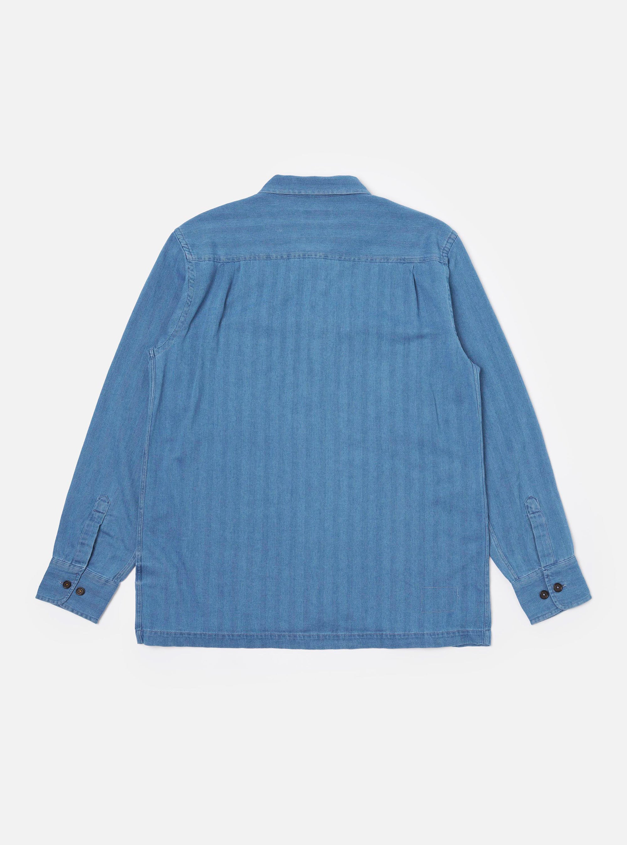 The Washed Indigo Recycled Herringbone Attendant Shirt is shown from the back as a flat lay against a plain white background. The image highlights the textured herringbone weave of the washed indigo fabric and the clean, simple construction of the garment's rear panel, featuring a straight hem.