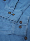 The Washed Indigo Recycled Herringbone Attendant Shirt is shown from the back as a flat lay against a plain white background. The image highlights the clean construction of the shirt's rear panel and the distinct V-shaped texture of the washed indigo herringbone fabric. The garment features a simple straight hem and a yoke across the shoulders.