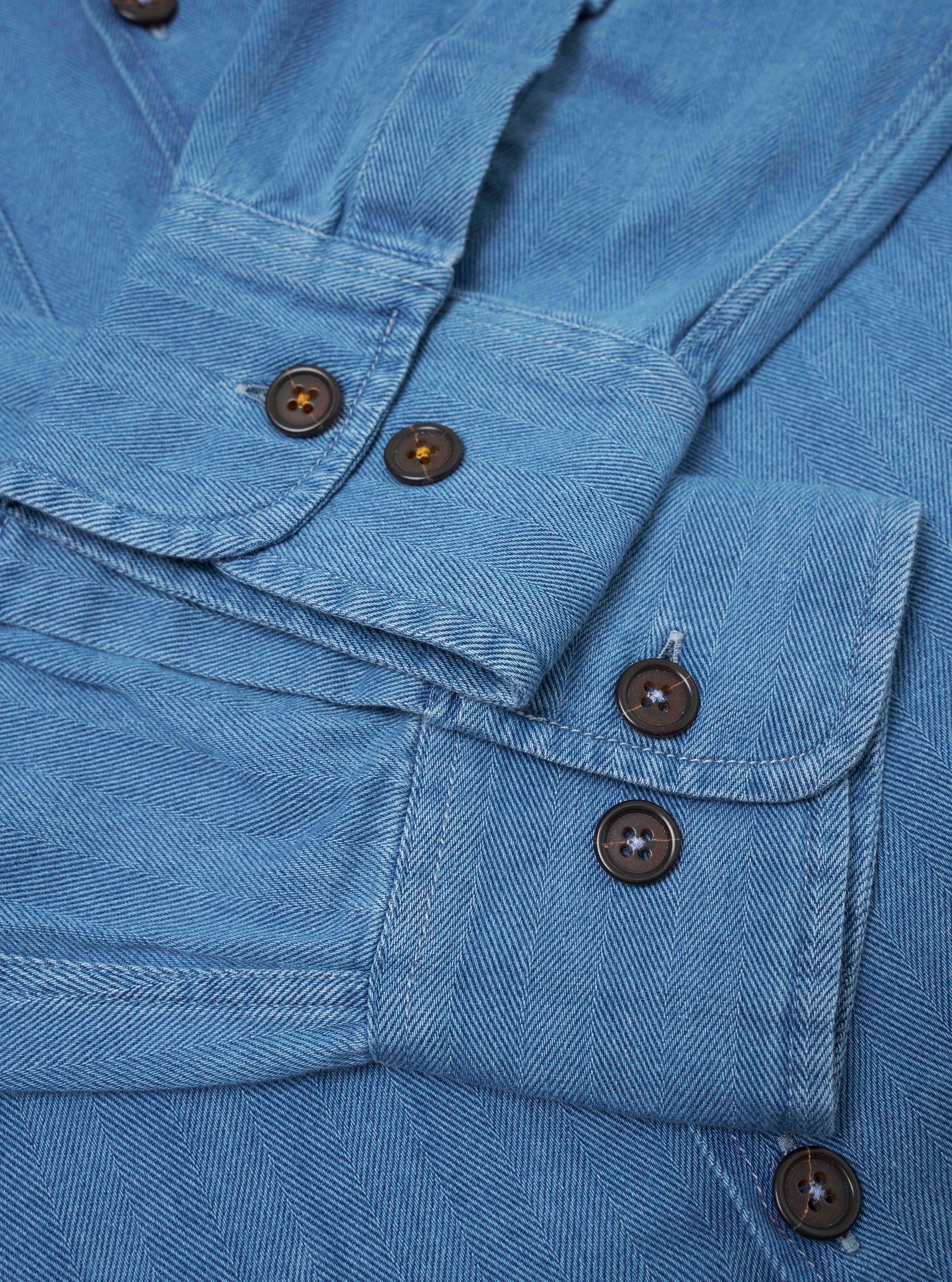 The Washed Indigo Recycled Herringbone Attendant Shirt is shown from the back as a flat lay against a plain white background. The image highlights the clean construction of the shirt's rear panel and the distinct V-shaped texture of the washed indigo herringbone fabric. The garment features a simple straight hem and a yoke across the shoulders.