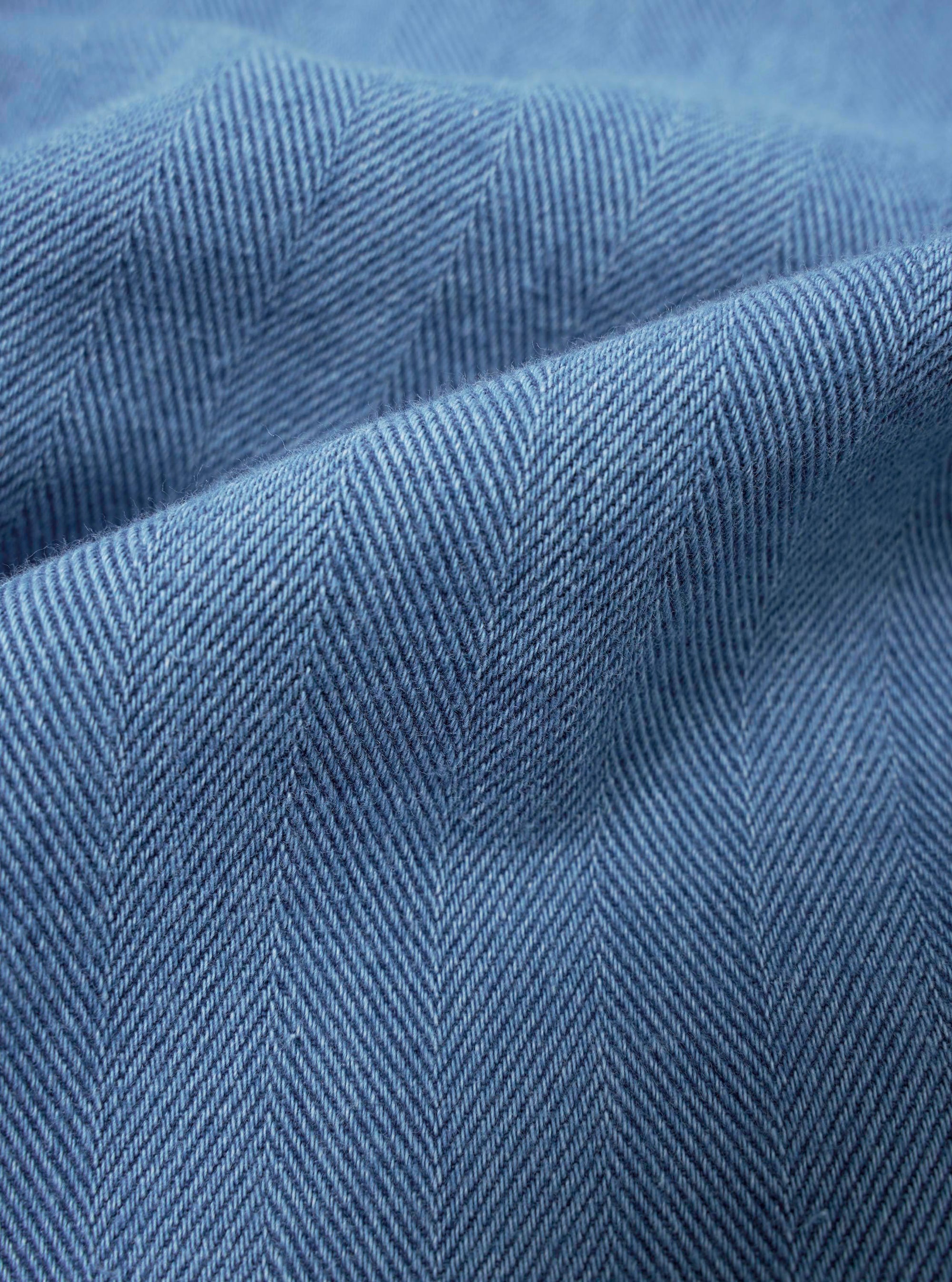 A close-up fabric swatch of the Washed Indigo Recycled Herringbone Attendant Shirt displays a deep indigo blue color with a distinct herringbone weave pattern. The texture of the recycled cotton material is clearly visible, highlighting the subtle tonal variations in the fabric.