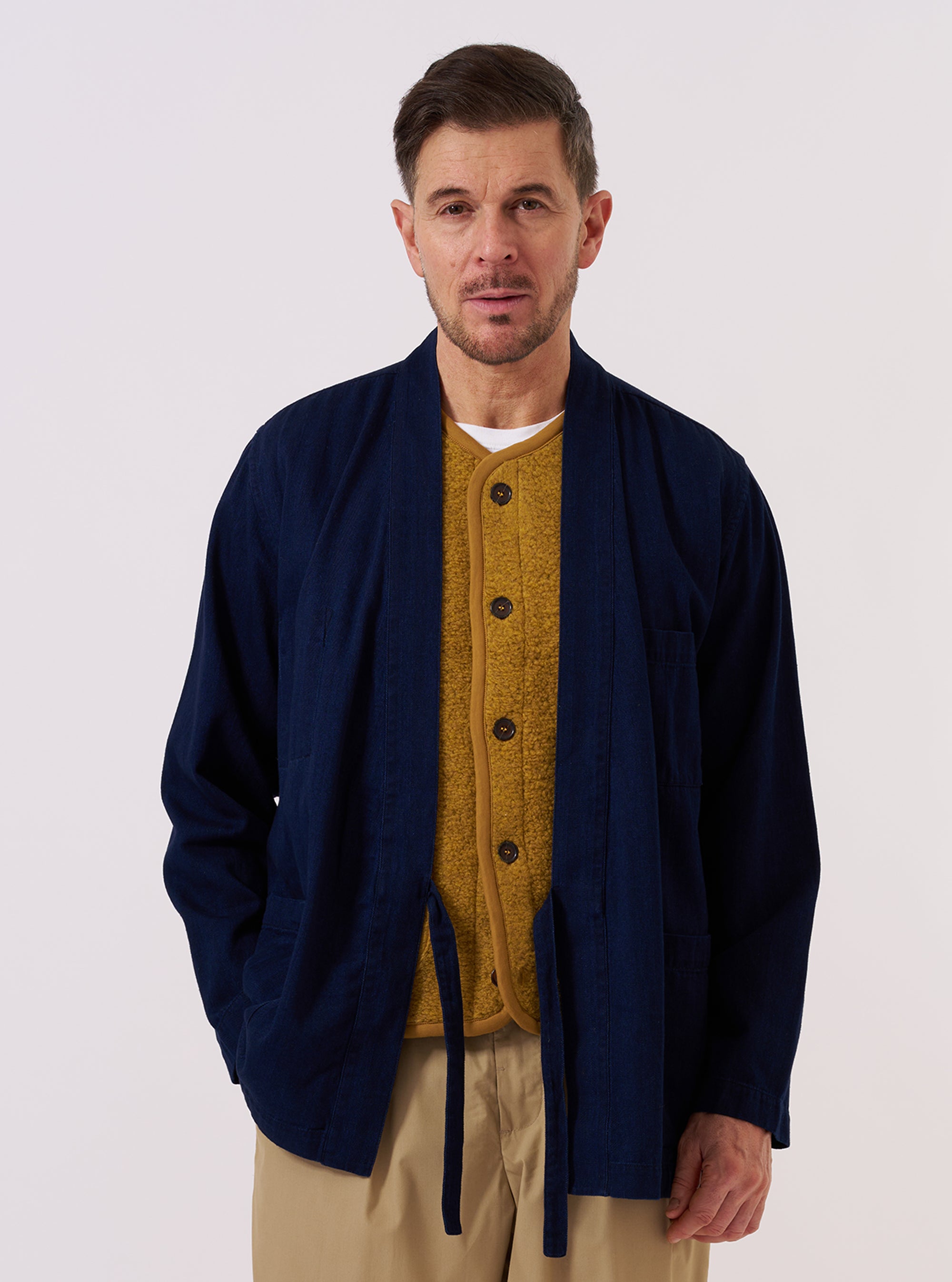 A model wears the Indigo Recycled Herringbone Kyoto Work Jacket while standing against a plain, neutral studio background. The jacket features a wrap-style front with a fabric side-tie closure, a straight hem, and a loose fit. Prominent details include the textured indigo herringbone denim fabric and two large patch pockets at the waist. The model is wearing the jacket over a light-colored top.