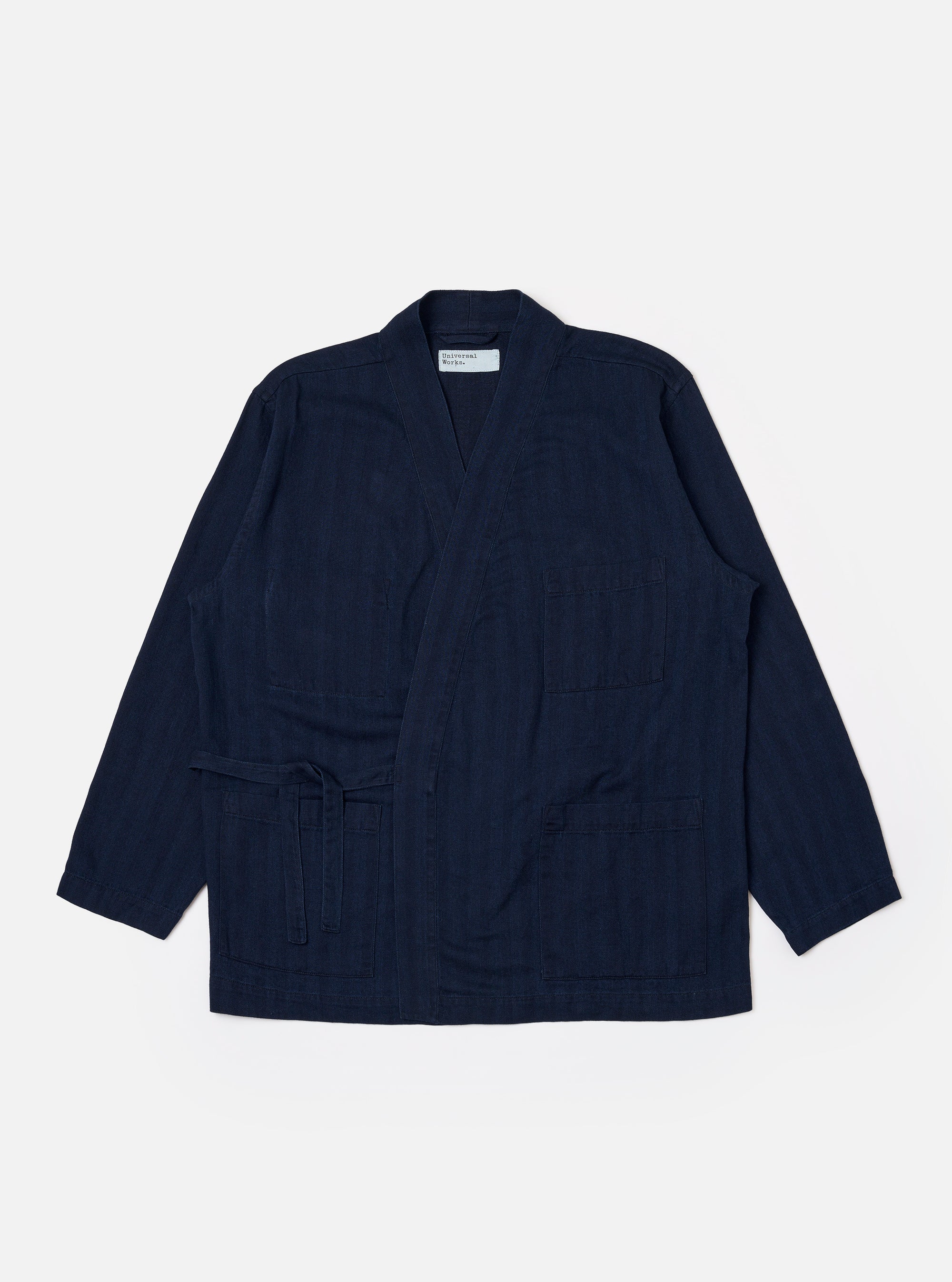 The Indigo Recycled Herringbone Kyoto Work Jacket is shown as a flat lay against a white background. The jacket features a collarless, wrap-style front with fabric ties, two large patch pockets at the waist, and one patch pocket on the left chest. The indigo-colored fabric has a visible herringbone weave texture.