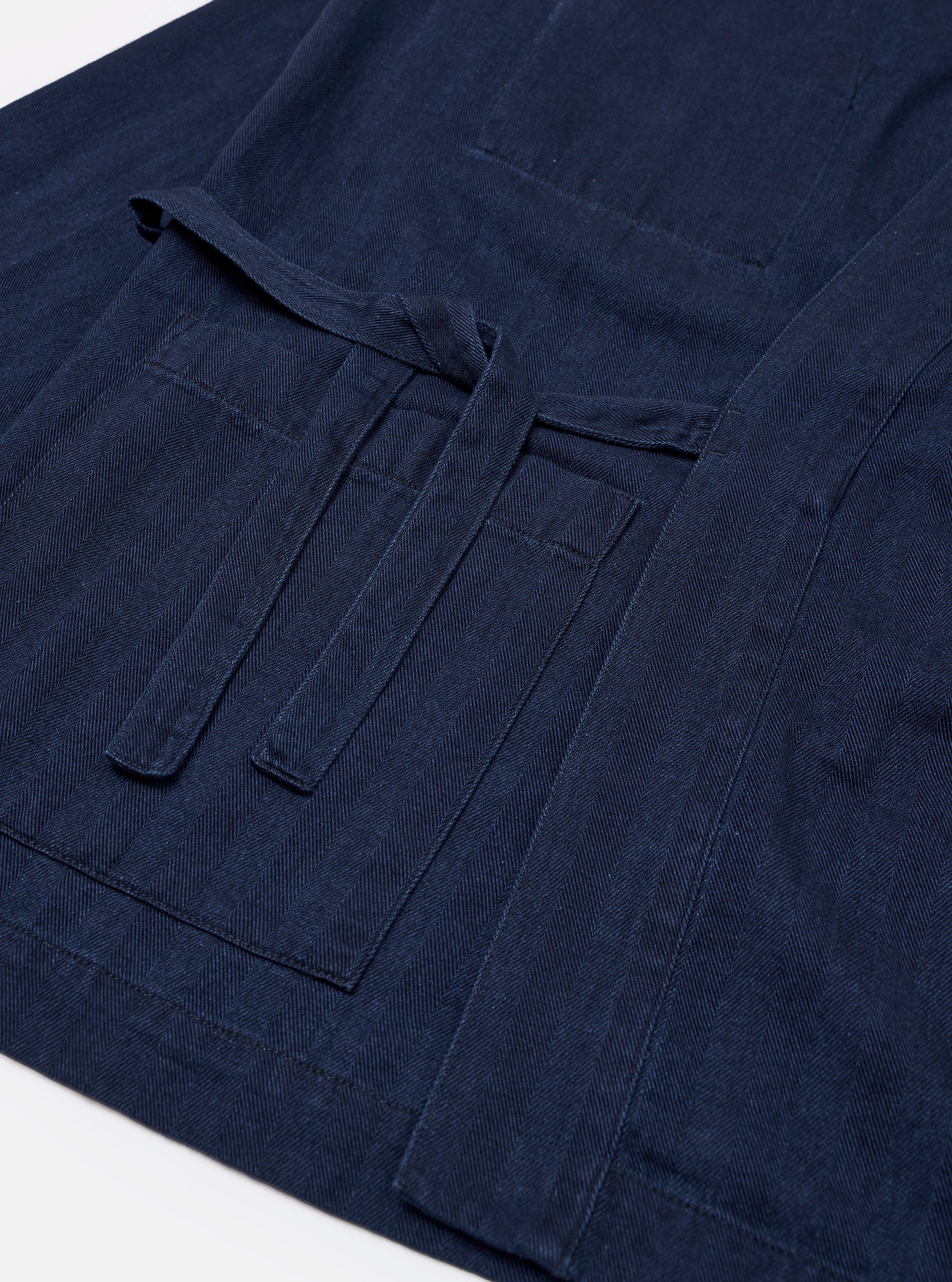 The Indigo Recycled Herringbone Kyoto Work Jacket is shown from the back as a flat lay against a plain white background. The image highlights the jacket's unstructured design, straight hem, and the textured indigo-washed herringbone cotton fabric.