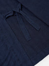 The Indigo Recycled Herringbone Kyoto Work Jacket is shown from the back as a flat lay against a plain white background. The image highlights the clean, straight-cut back panel and the wrap-around ties that extend from the sides. The washed indigo fabric features a distinct herringbone weave throughout.