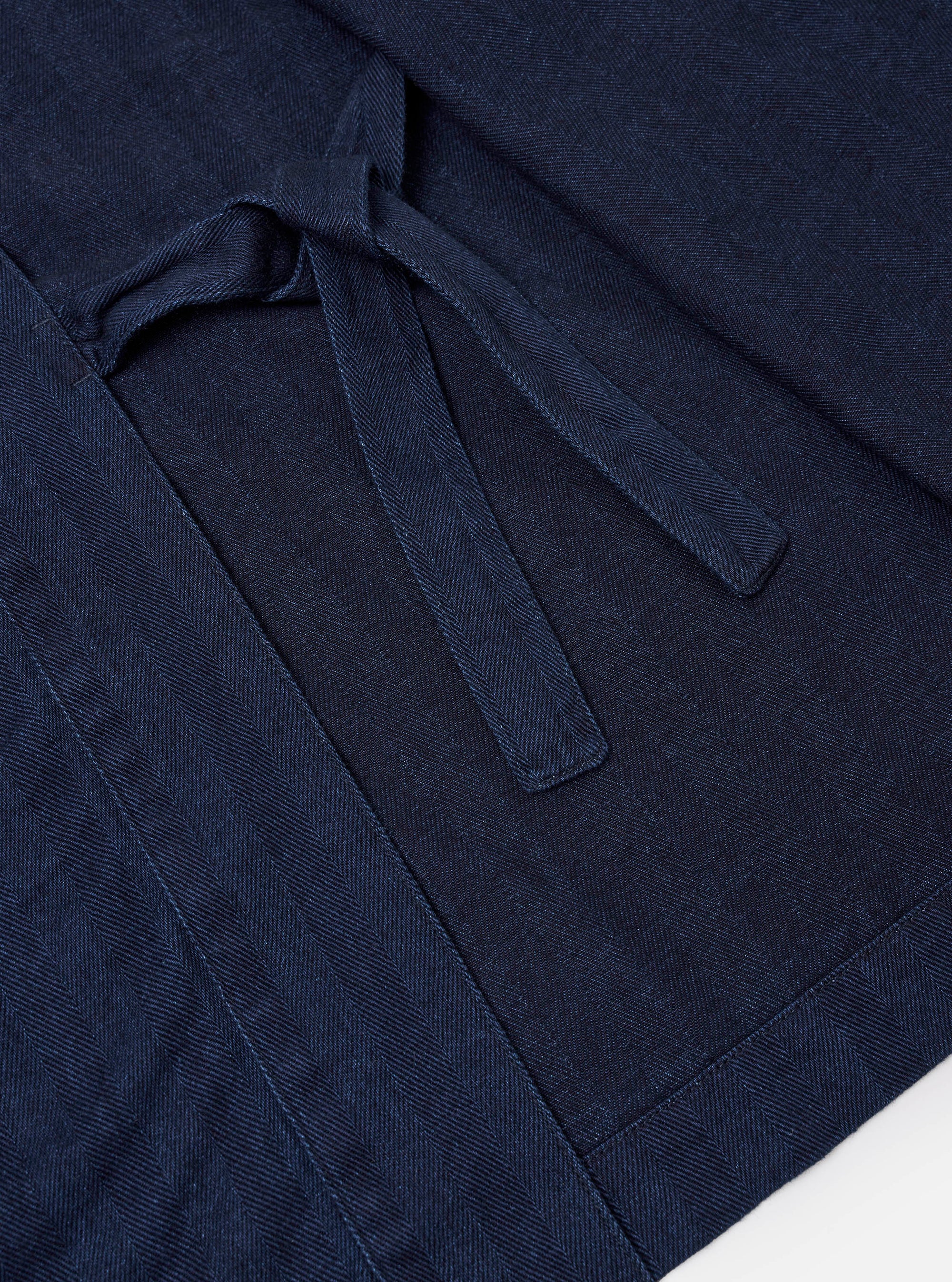 The Indigo Recycled Herringbone Kyoto Work Jacket is shown from the back as a flat lay against a plain white background. The image highlights the clean, straight-cut back panel and the wrap-around ties that extend from the sides. The washed indigo fabric features a distinct herringbone weave throughout.