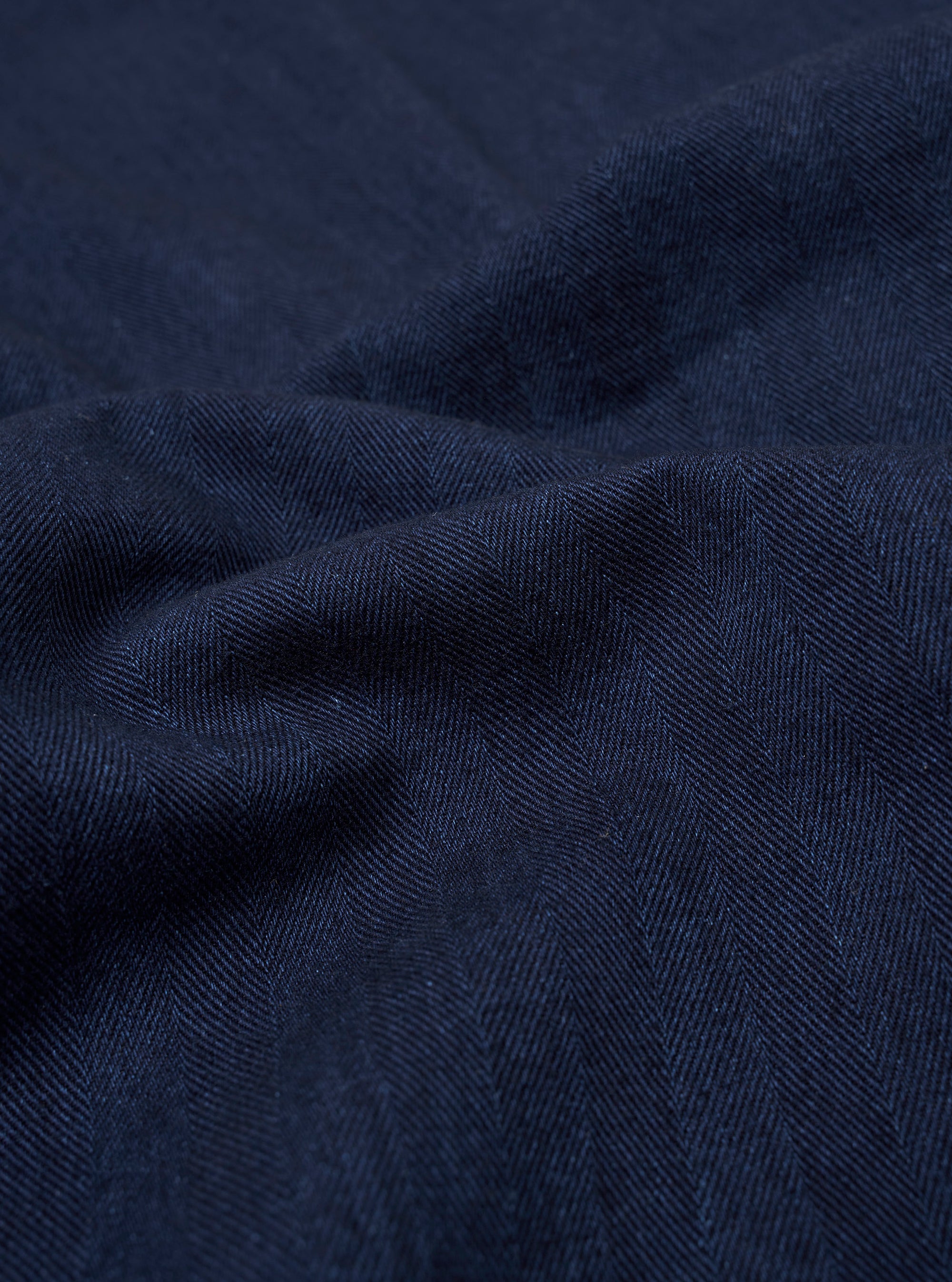 A close-up swatch of the Indigo Recycled Herringbone Kyoto Work Jacket highlights the deep indigo blue color and the distinct V-shaped herringbone weave of the fabric. The textured material fills the entire frame.
