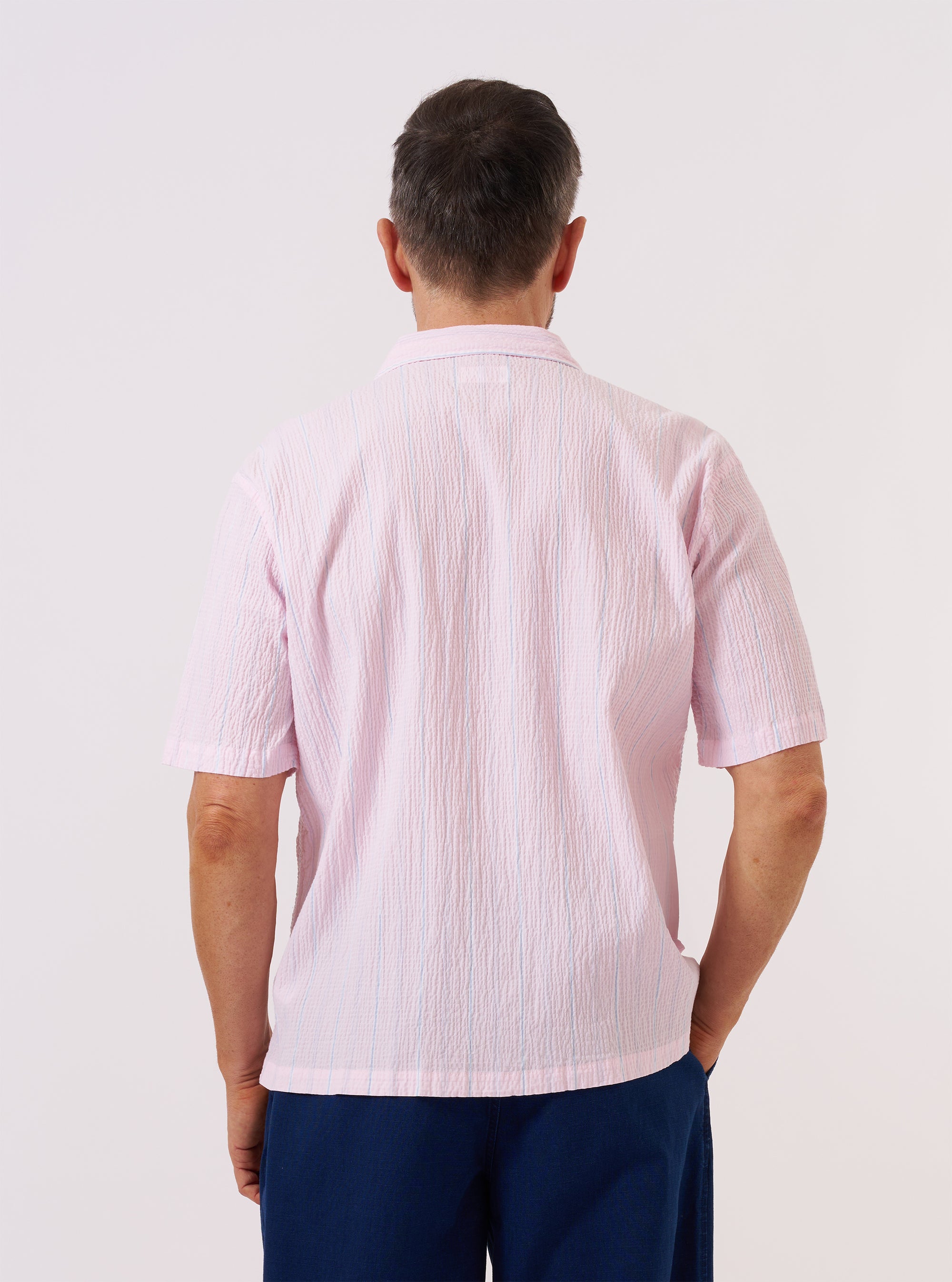 A model wears the Pink Lily Stripe Road Shirt against a plain, light-colored background. The short-sleeve shirt features a vertical pink and white pinstripe pattern on a textured seersucker fabric, a one-piece camp collar worn open, and a single chest patch pocket. The garment is shown with a regular fit and a straight hem.