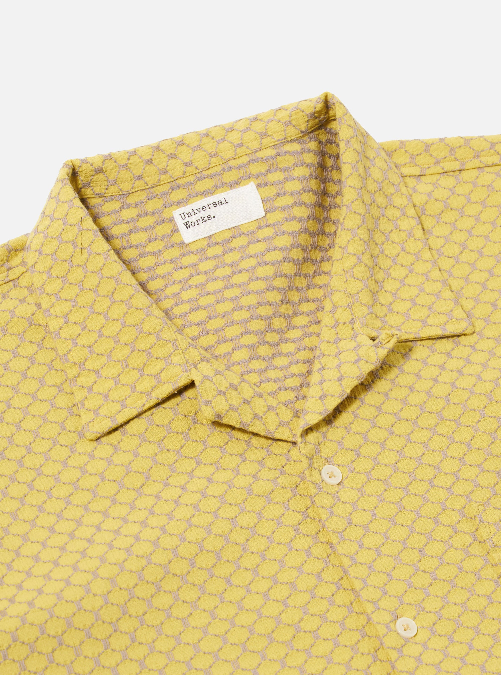 A model wears the Yellow Mykonos Cotton Road Shirt against a plain, light-colored background. The shirt features a relaxed camp collar, a button-down front with tonal buttons, and a single chest patch pocket. The fabric displays a vibrant yellow and ochre patterned design, characteristic of the Mykonos cotton weave. The model is shown from the waist up, highlighting the garment's fit and textural detail.