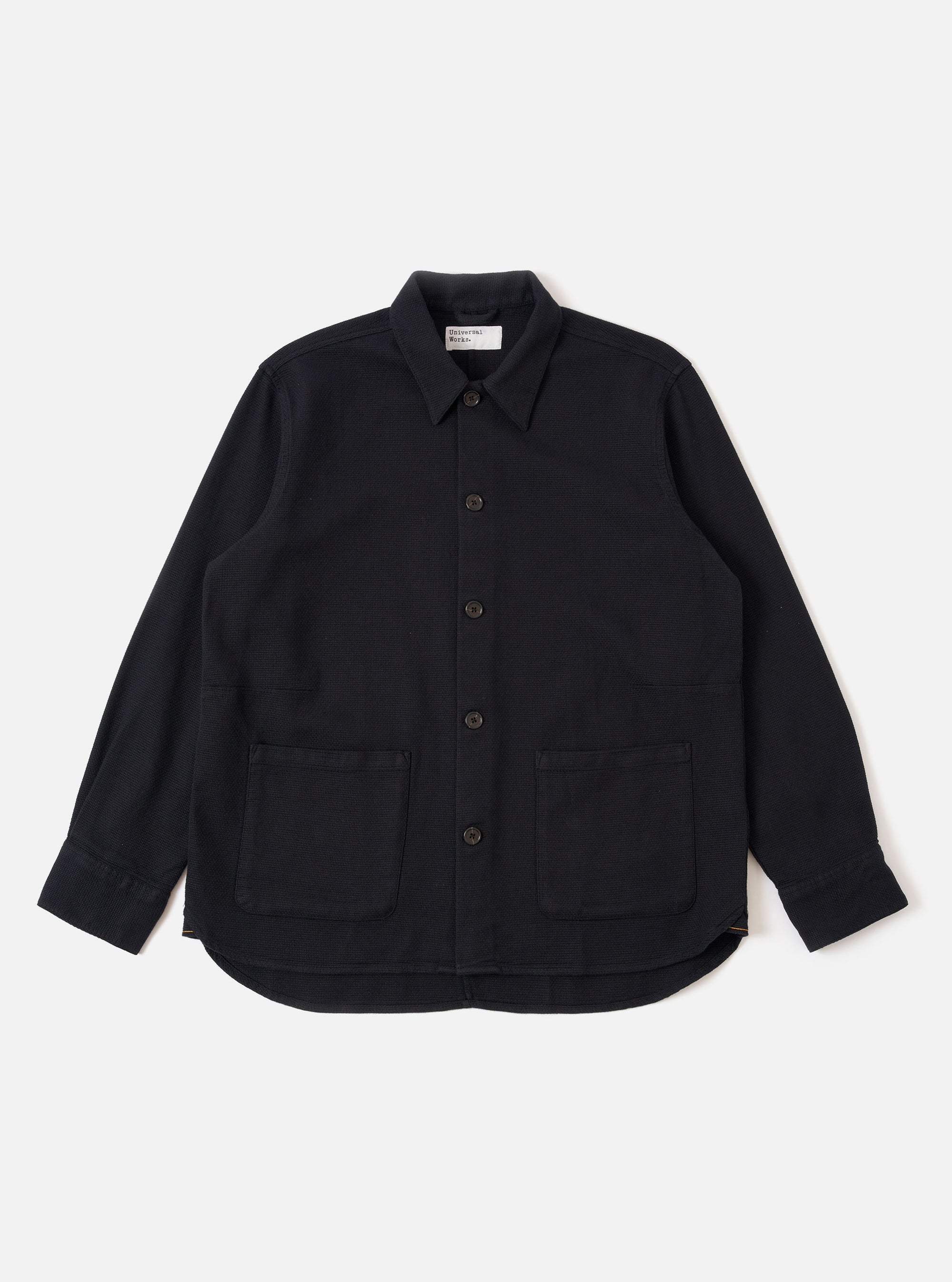The Black Kendal Cotton Travail Overshirt is shown as a flat lay against a white background. The overshirt features a one-piece collar, a central button-down closure, and two large patch pockets at the waist. The black cotton fabric has a smooth texture, and the garment is finished with a curved hem.