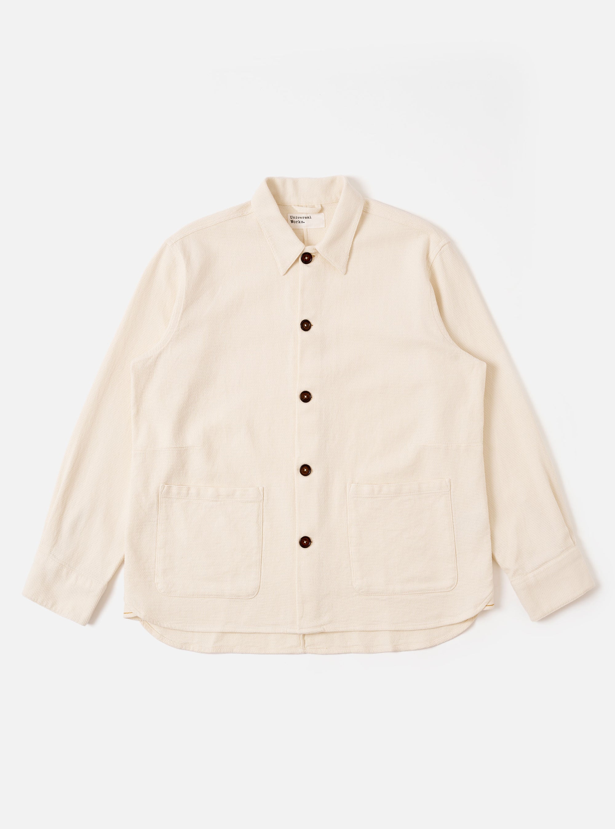 The Ecru Kendal Cotton Travail Overshirt is shown as a flat lay against a white background. The overshirt features a one-piece collar, a central button-down closure, and two large patch pockets at the waist. The garment is made from a textured ecru cotton fabric and has a curved hem with a visible contrast stitch insert.