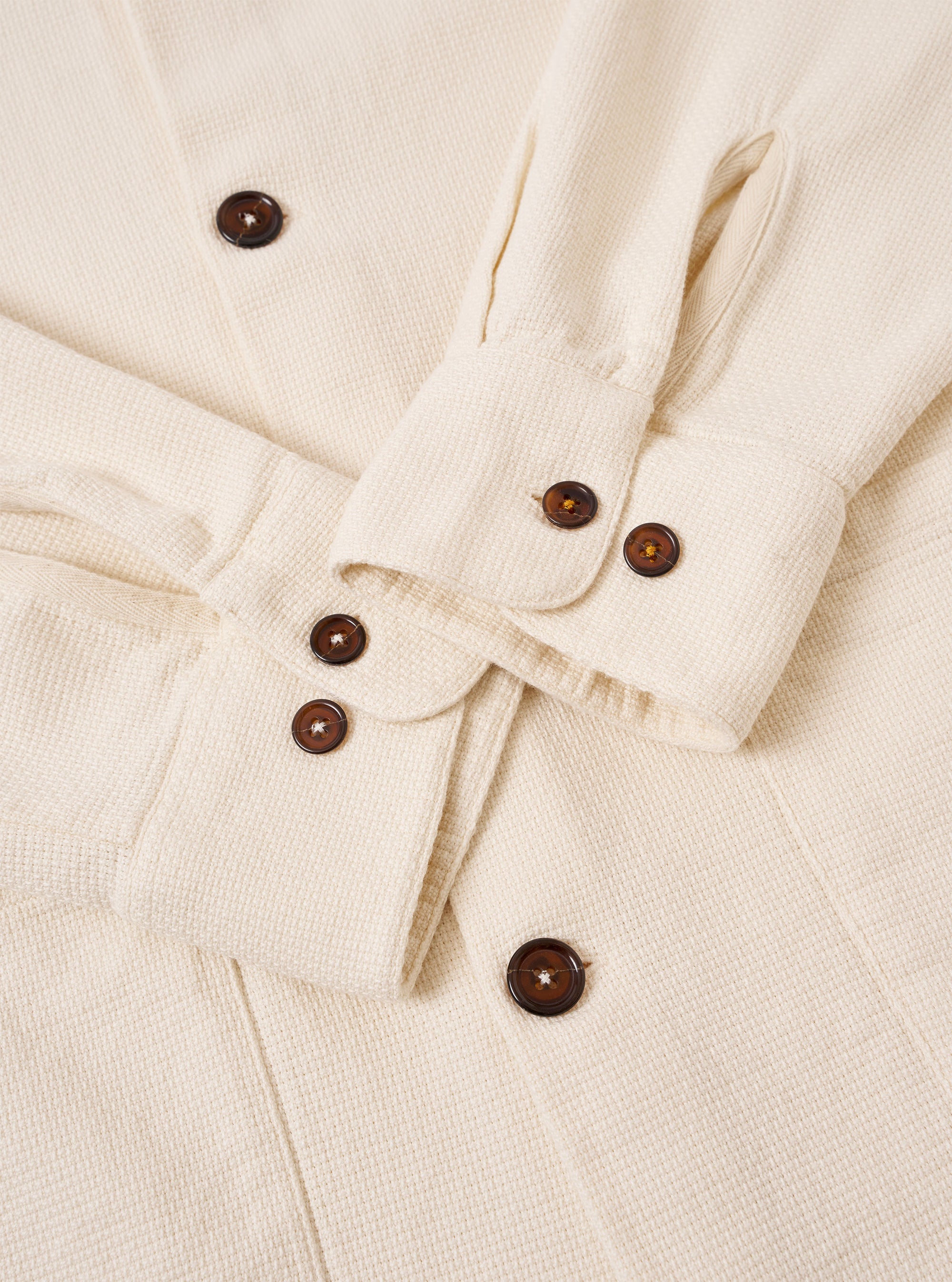 The Ecru Kendal Cotton Travail Overshirt is shown in a detail view as a flat lay against a plain white background. The image focuses on the lower corner of the garment, highlighting the curved shirt-like hem with a contrast stitch insert and the corner of a large patch pocket. The ecru-colored cotton fabric features a visible, textured open weave.