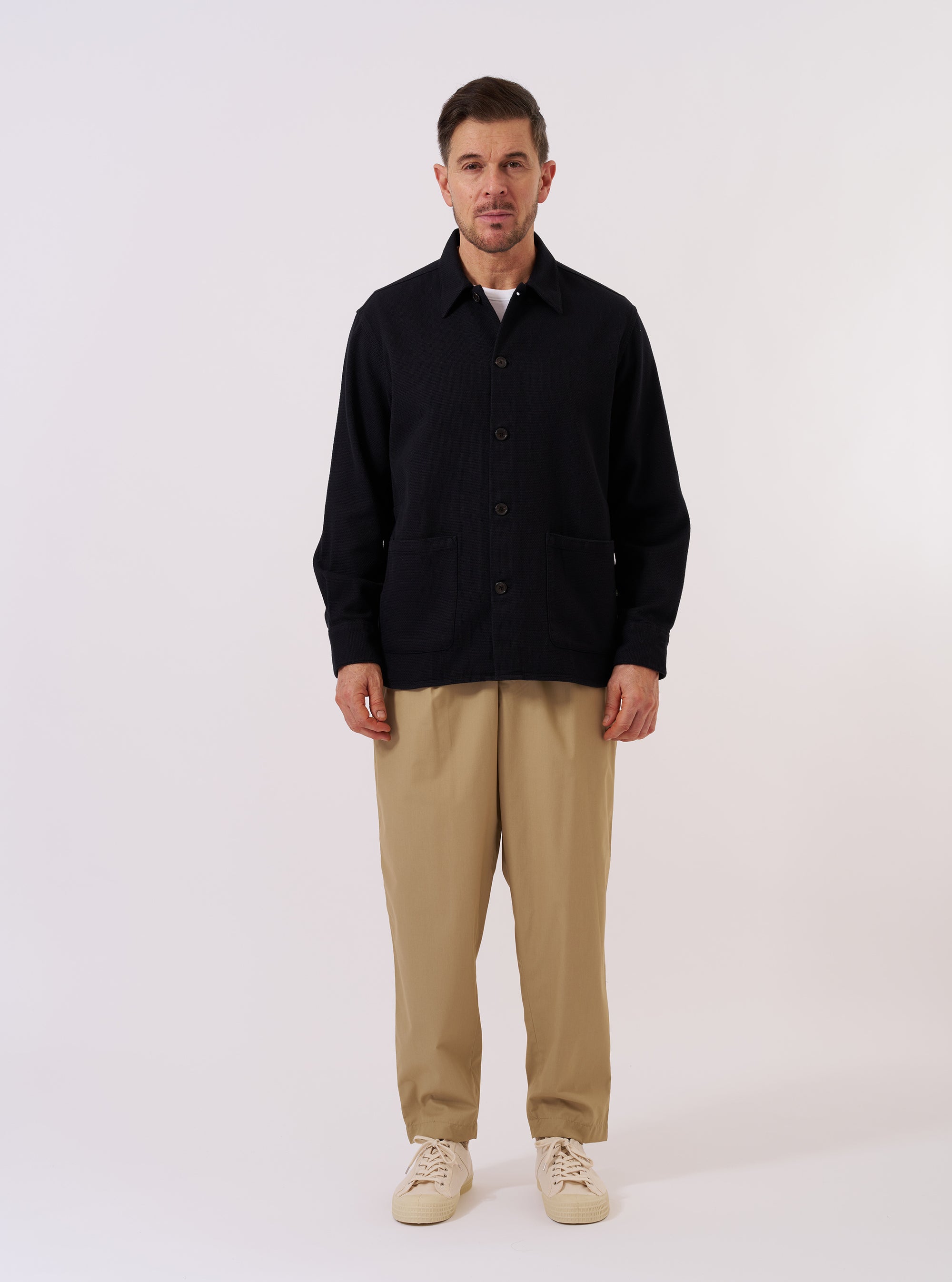 The Black Kendal Cotton Travail Overshirt is presented as a flat lay against a plain white background. The overshirt features a one-piece collar, a central button-down closure, and two large patch pockets at the waist. The black cotton fabric has a distinct textured appearance. The garment is finished with a curved hem and subtle chest darts.