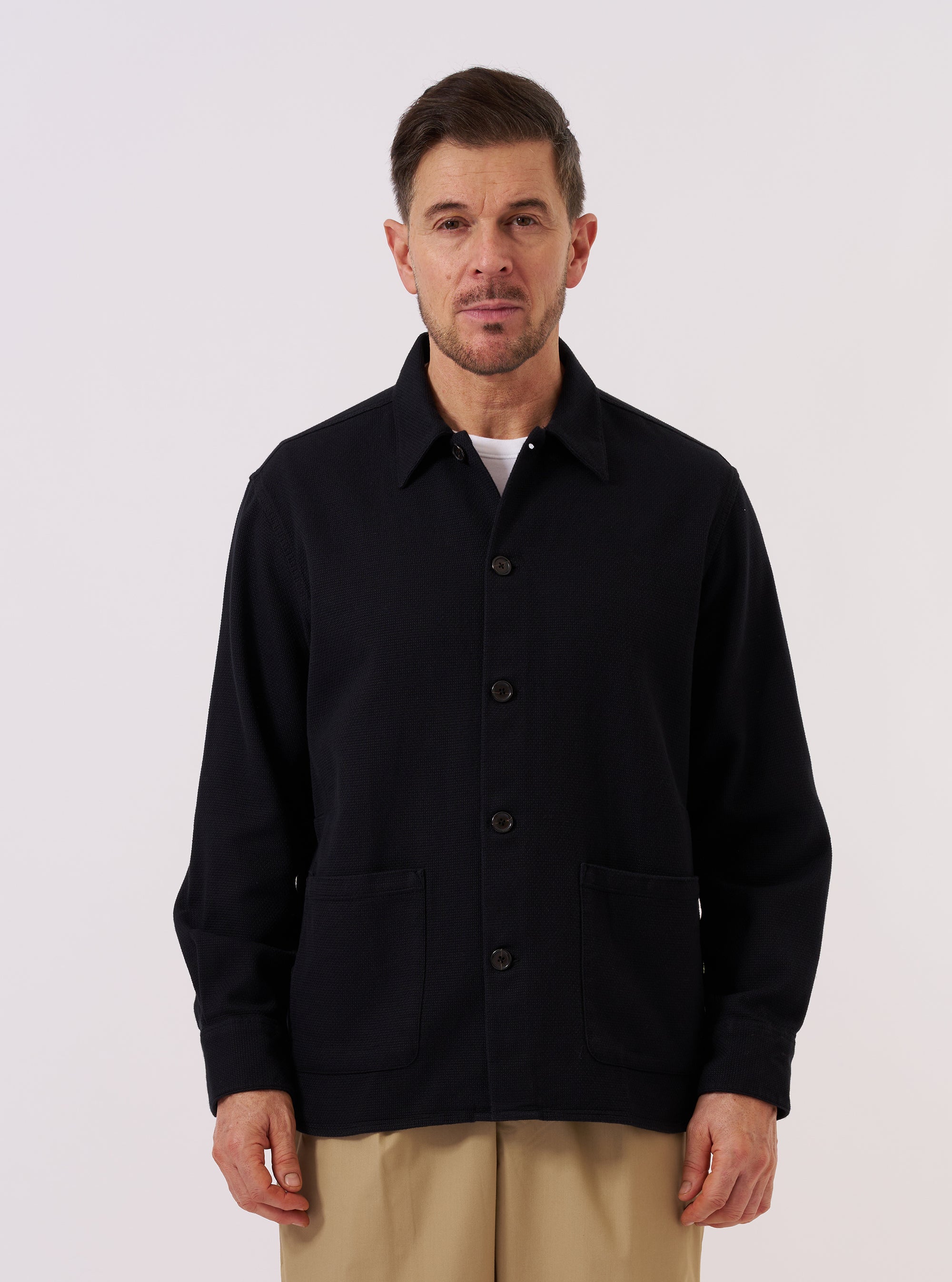 A model wears the Black Kendal Cotton Travail Overshirt over a light-colored t-shirt against a plain white background. The overshirt is shown from the front, highlighting its loose, unstructured fit, one-piece collar, and central button-down closure. Two large patch pockets are visible at the waist, and the black cotton fabric features a clean, matte finish.