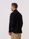 A model wears the Black Kendal Cotton Travail Overshirt, shown from a rear three-quarter angle against a plain white background. The image highlights the garment's loose, unstructured fit and its distinctive curved, shirt-like hem. The black cotton fabric has a smooth finish, and the one-piece collar is visible at the neck.