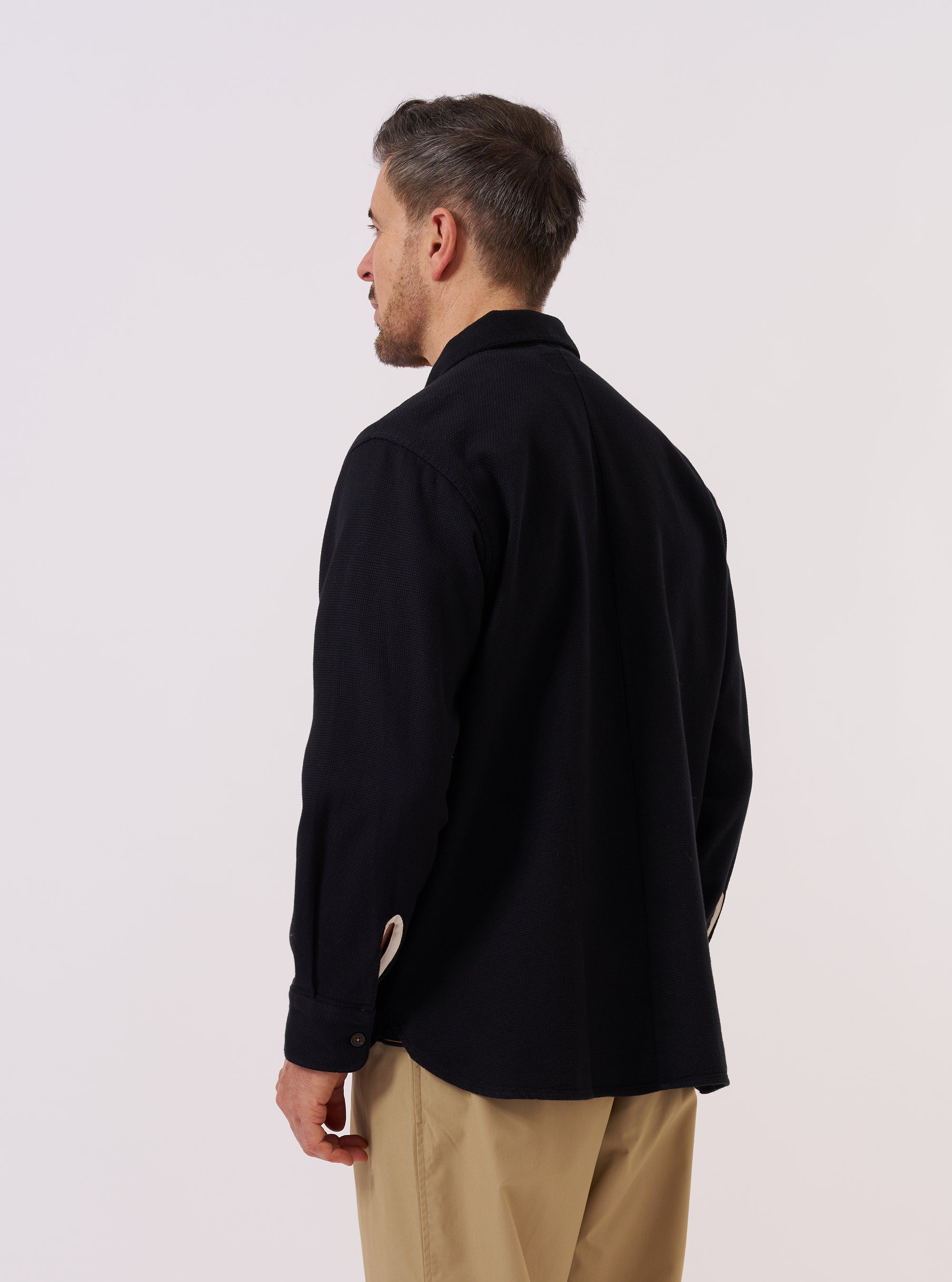 A model wears the Black Kendal Cotton Travail Overshirt, shown from a rear three-quarter angle against a plain white background. The image highlights the garment's loose, unstructured fit and its distinctive curved, shirt-like hem. The black cotton fabric has a smooth finish, and the one-piece collar is visible at the neck.