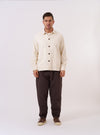 The Ecru Kendal Cotton Travail Overshirt is shown as a flat lay against a plain white background. The overshirt features a classic one-piece collar, a central button-down closure, and two large patch pockets at the waist. The textured ecru cotton fabric has a visible open weave, and the garment is finished with a curved hem and adjustable button cuffs.