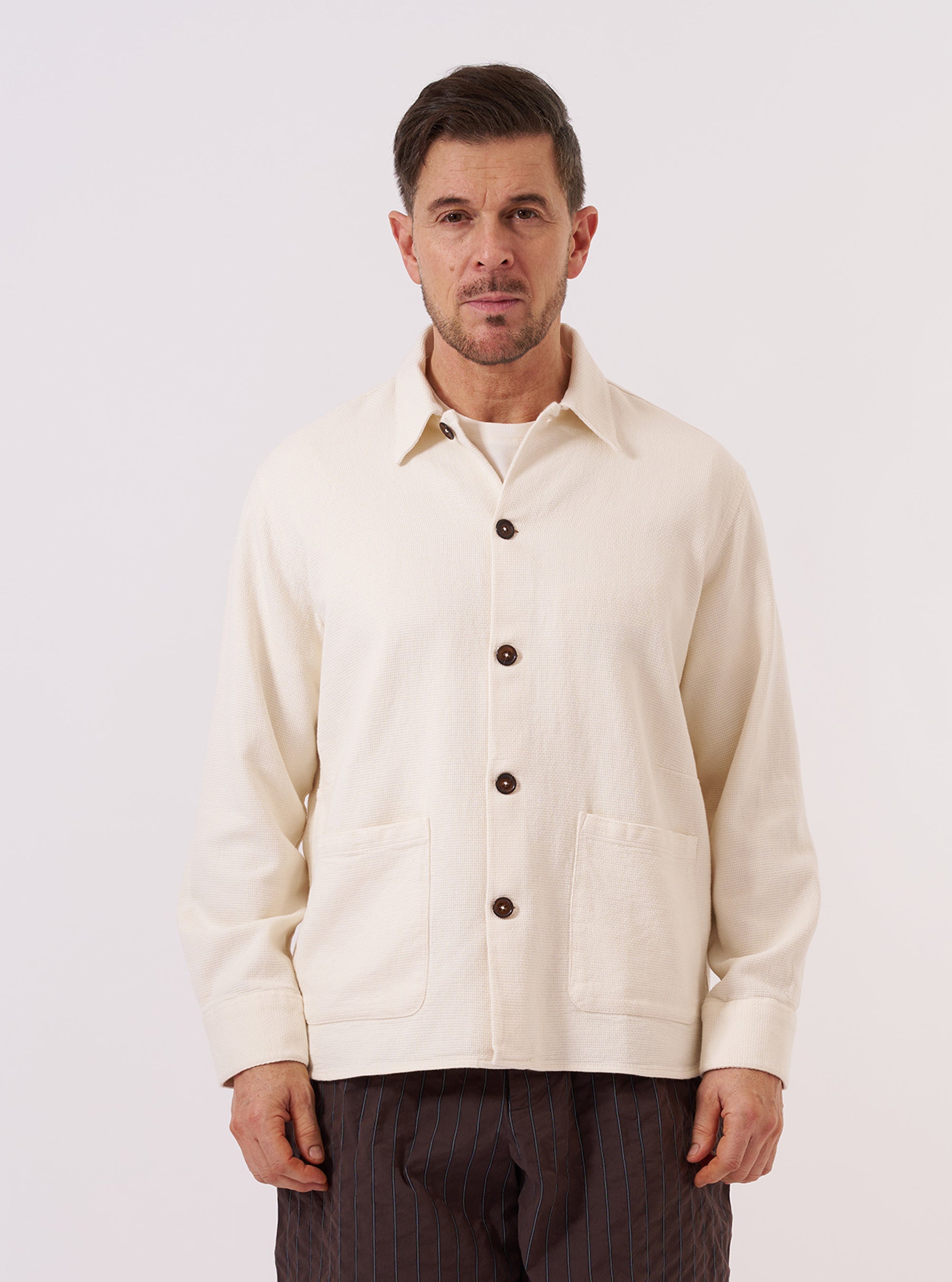 A model wears the Ecru Kendal Cotton Travail Overshirt against a plain, neutral background. The overshirt features a one-piece collar, a central button-down closure, and two large patch pockets at the waist. The garment is made from an off-white ecru cotton fabric and has a loose, unstructured fit.