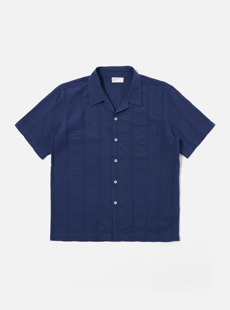 The Blue Maui Cotton Road Shirt is shown as a flat lay against a white background. The short-sleeve shirt features a vertical blue and white stripe pattern, a one-piece camp collar with a loop-button closure at the neck, and a single large patch pocket on the left chest. It has a straight hem and