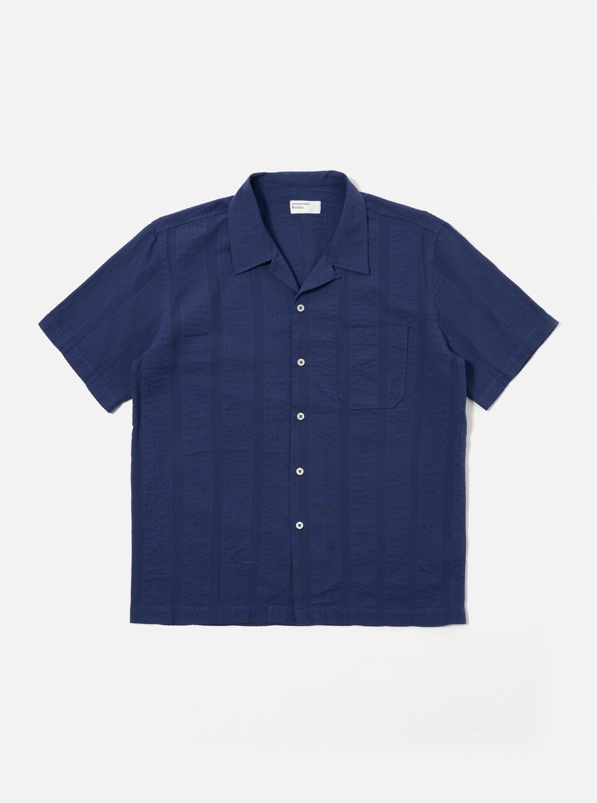 The Blue Maui Cotton Road Shirt is shown as a flat lay against a white background. The short-sleeve shirt features a vertical blue and white stripe pattern, a one-piece camp collar with a loop-button closure at the neck, and a single large patch pocket on the left chest. It has a straight hem and