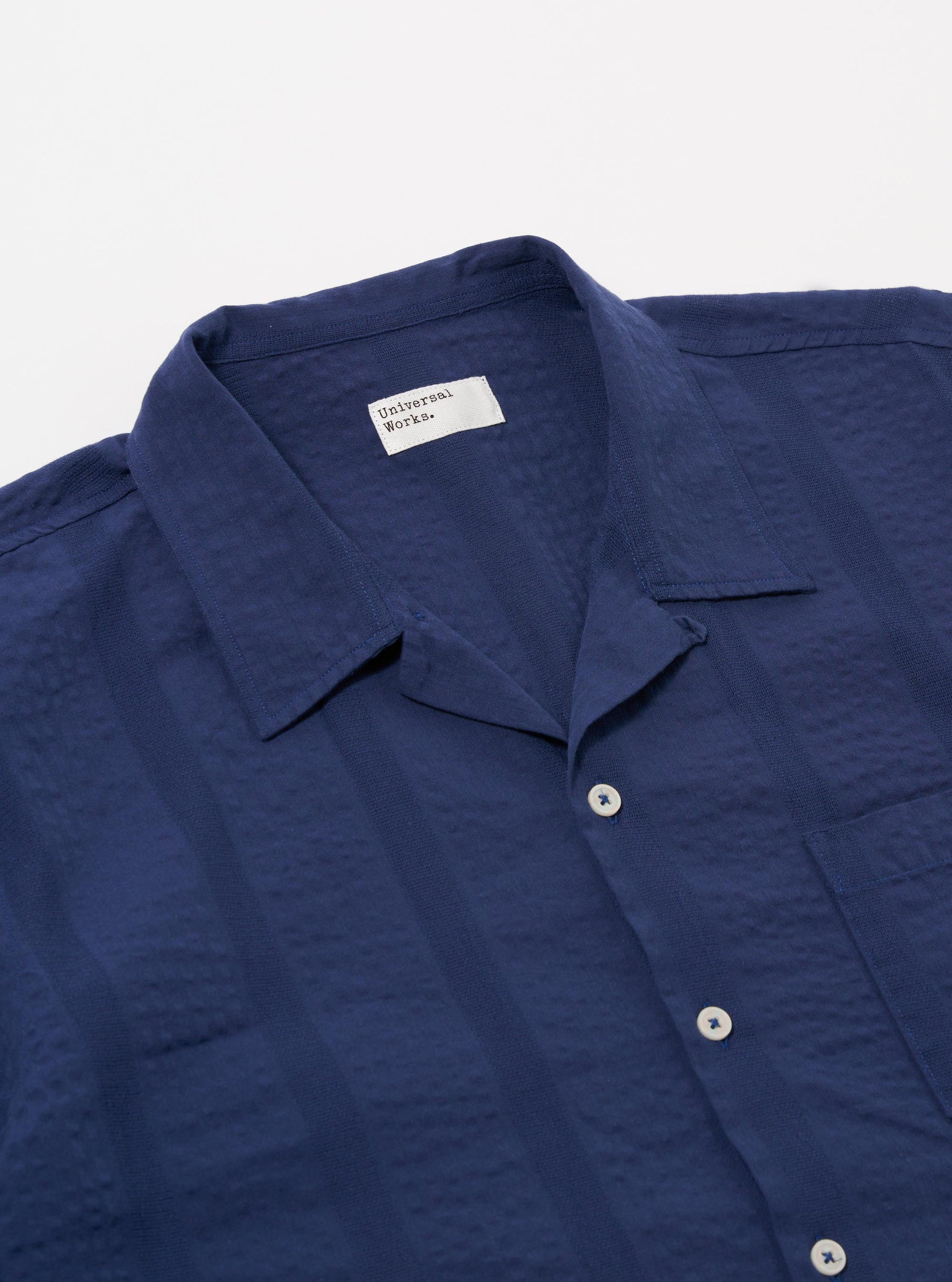 A model wears the Blue Maui Cotton Road Shirt over a white t-shirt against a plain white background. The short-sleeved shirt features a vertical blue and white stripe pattern, a camp collar, and a single chest pocket. The model is shown from the chest up, with the shirt left unbuttoned.