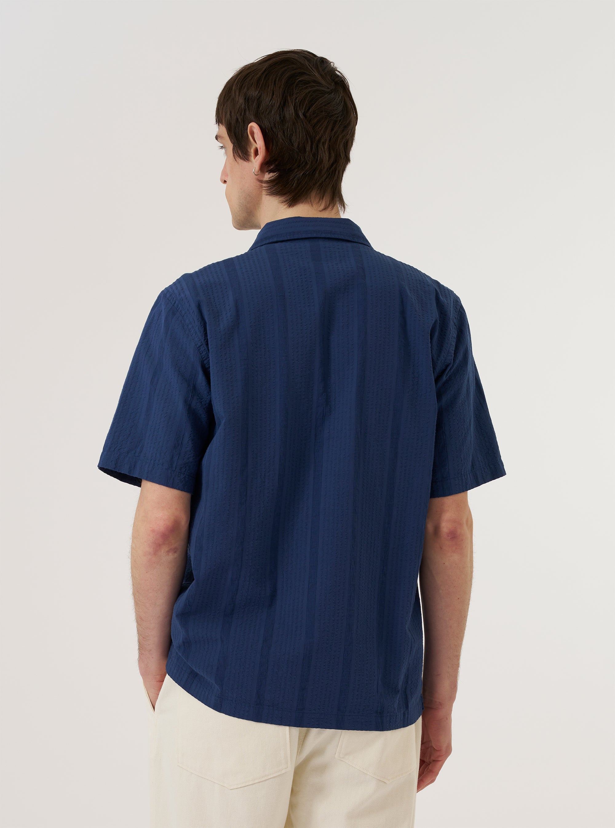 The Blue Maui Cotton Road Shirt is worn by a model against a plain white background. The image is a rear view showing the model from the head down to the waist. The shirt features a vertical blue and white stripe pattern, a straight hem, and a single-piece collar with a small loop detail at the neck.