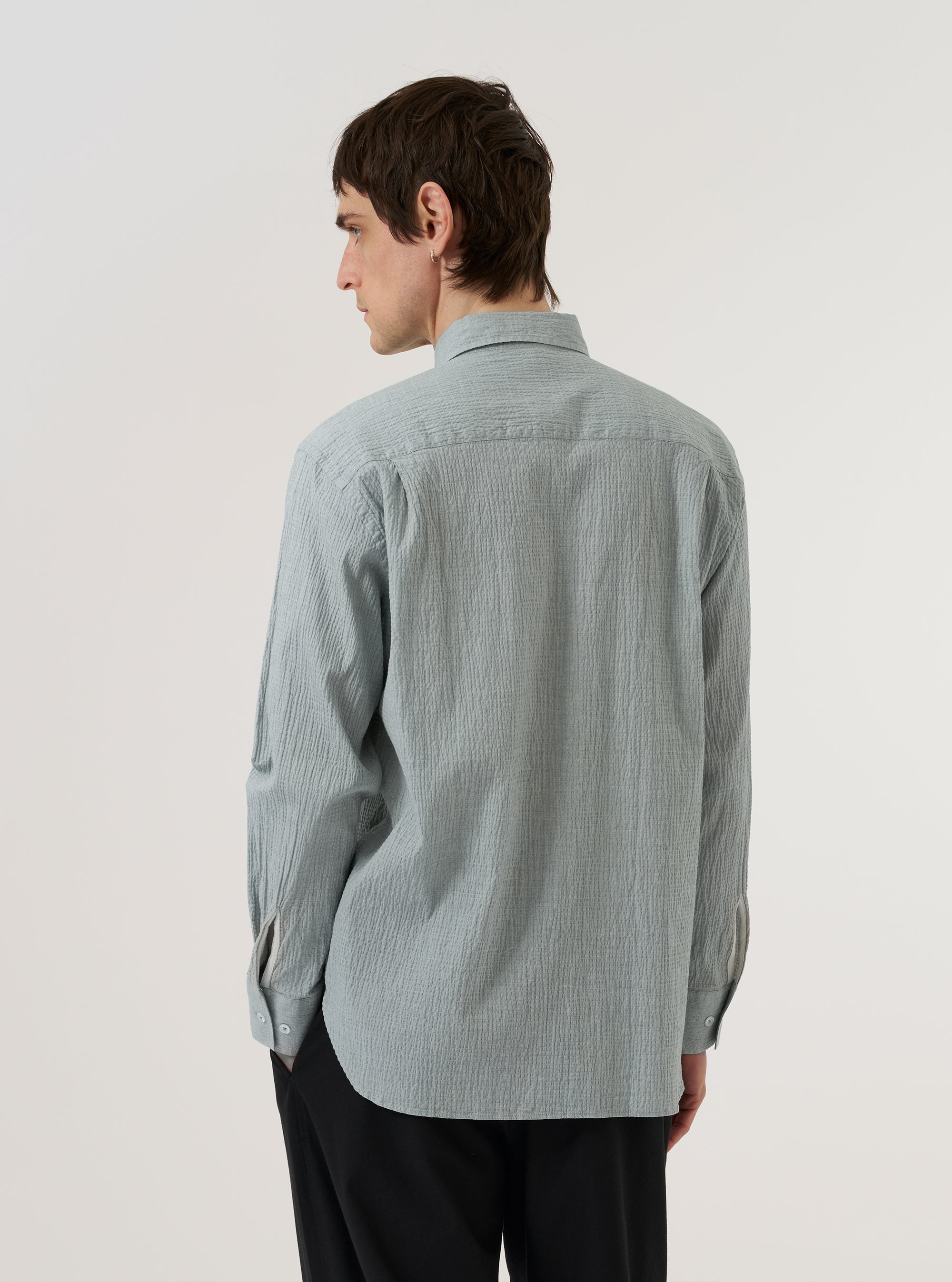 A model wears the Green Seersucker Square Pocket Shirt against a plain white background, shown from a rear three-quarter angle. The shirt features a relaxed fit, short sleeves, and a straight hem. The light green fabric has a distinct puckered seersucker texture throughout.