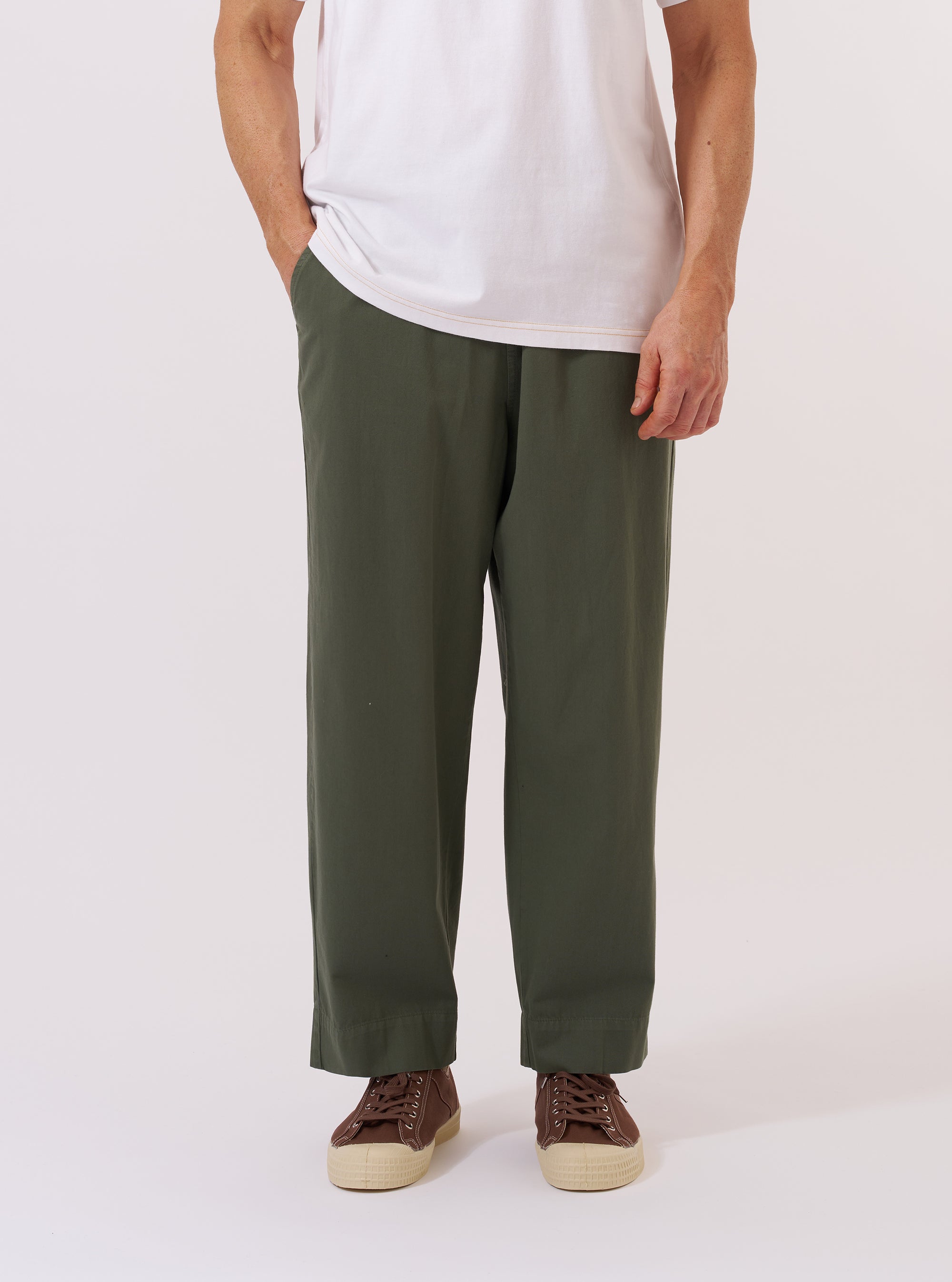 A model wears the Olive Kelly Cotton Judo Pant against a plain, neutral background. The trousers feature a relaxed, loose fit and are shown from the waist down, highlighting the comfortable drawstring waistband and the textured olive green cotton fabric. The straight-leg cut and functional design of the judo-style pants are the prominent features.
