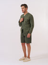 The Olive Kelly Cotton Glider Jacket is shown as a flat lay against a plain white background. The jacket features a collarless design with bound edges, a central two-way zip closure, and four large 3D bellows pockets on the front. The fabric is a solid olive green cotton.