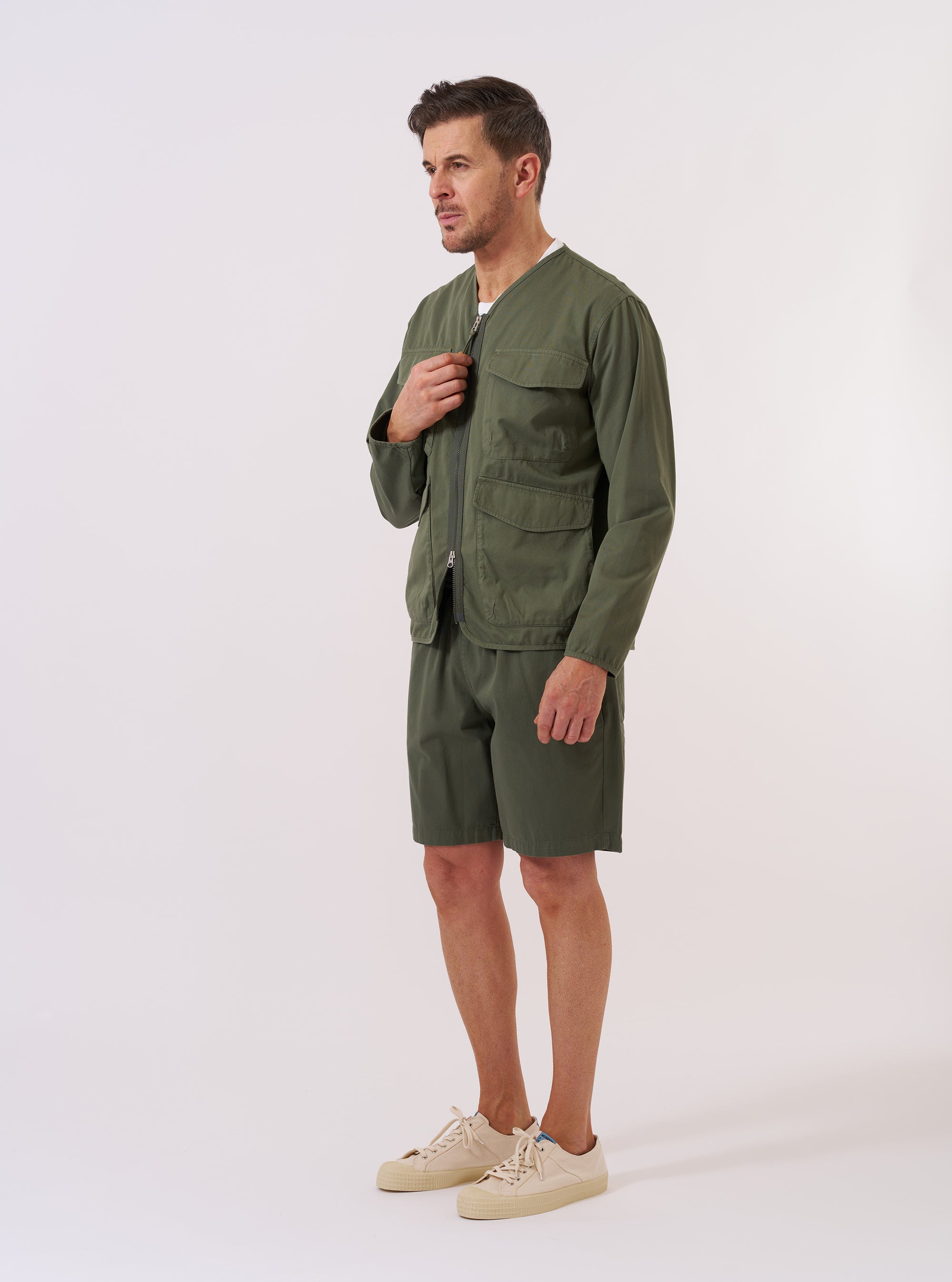 The Olive Kelly Cotton Glider Jacket is shown as a flat lay against a plain white background. The jacket features a collarless design with bound edges, a central two-way zip closure, and four large 3D bellows pockets on the front. The fabric is a solid olive green cotton.