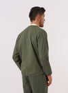 A model wears the Olive Kelly Cotton Glider Jacket over a white t-shirt, shown from the rear three-quarter angle against a plain white background. The image highlights the jacket's loose fit, simple straight hem, and the smooth texture of the olive green cotton fabric. One of the large front patch pockets is partially visible from the side.
