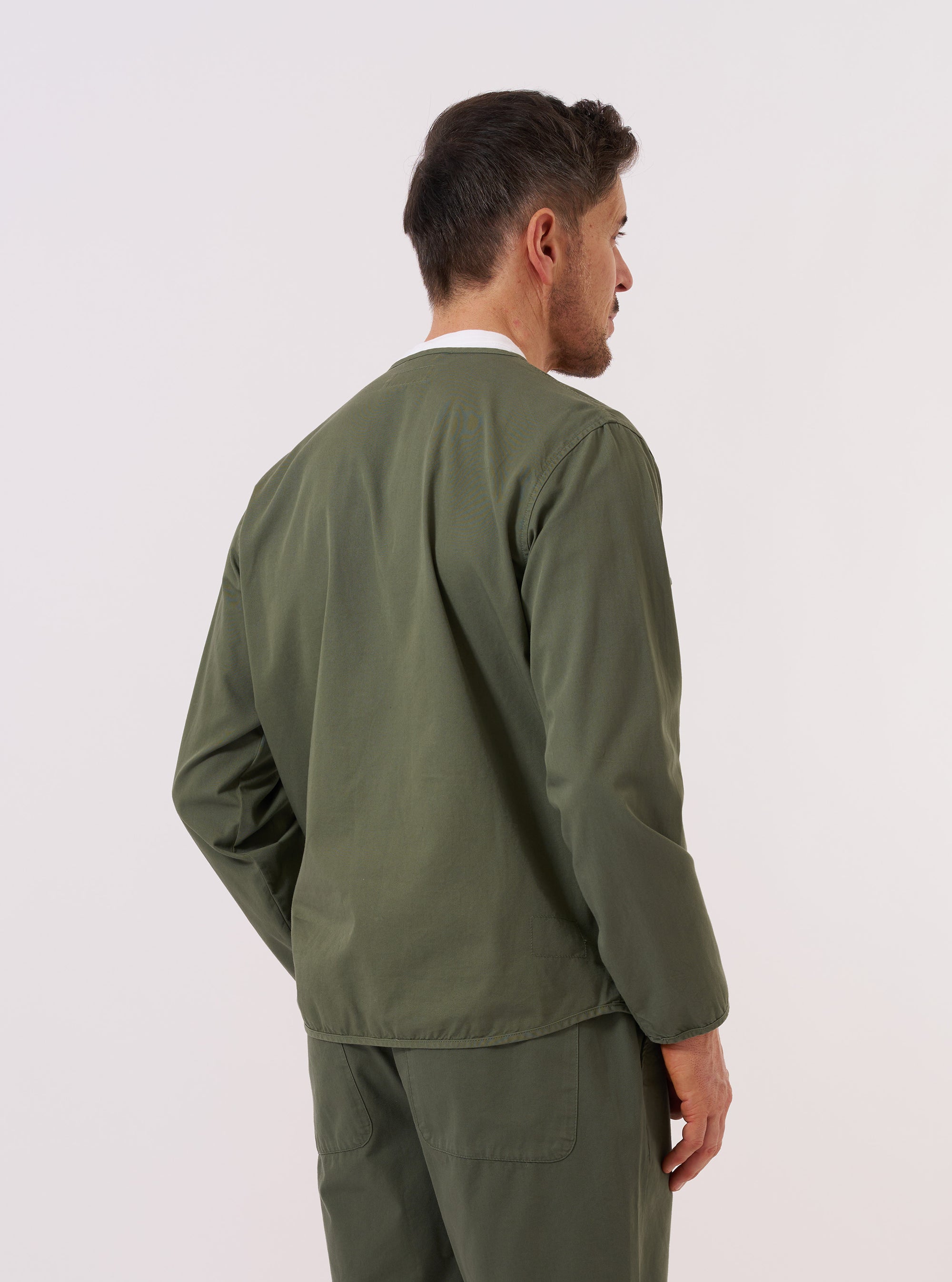 A model wears the Olive Kelly Cotton Glider Jacket over a white t-shirt, shown from the rear three-quarter angle against a plain white background. The image highlights the jacket's loose fit, simple straight hem, and the smooth texture of the olive green cotton fabric. One of the large front patch pockets is partially visible from the side.