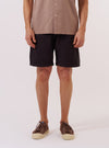 A model wears the Licorice Lyo Linen Mix Simple Short against a plain white background. The shorts feature a relaxed fit, an elasticated waistband with a drawstring, and side pockets. The fabric has a textured, lightweight appearance in a dark brown licorice color. The model is shown from the waist down, paired with a light-colored shirt and dark sandals.