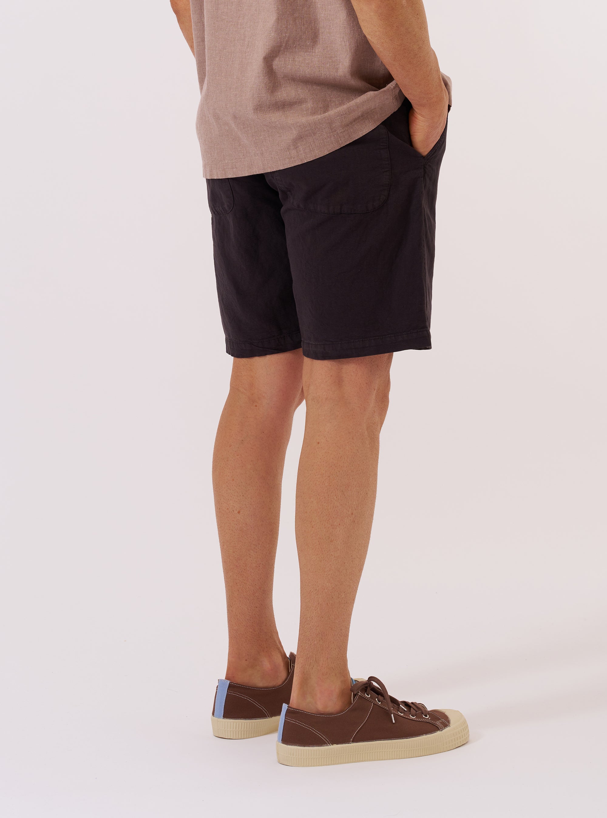 A model is wearing the Licorice Lyo Linen Mix Simple Short, shown from the waist down from a rear perspective against a plain white background. The dark brown shorts feature a relaxed fit and a single patch pocket on the back.