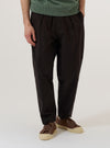 A model wears the Licorice Lyo Linen Mix Oxford Pant, shown from the waist down against a plain, neutral studio background. These loose-fit trousers in a deep brown licorice color feature a pleated front, a drawstring at the elasticated waistband, and side pockets. The model is wearing the pants with a tucked-in shirt and dark shoes, highlighting the relaxed, wide-leg silhouette.