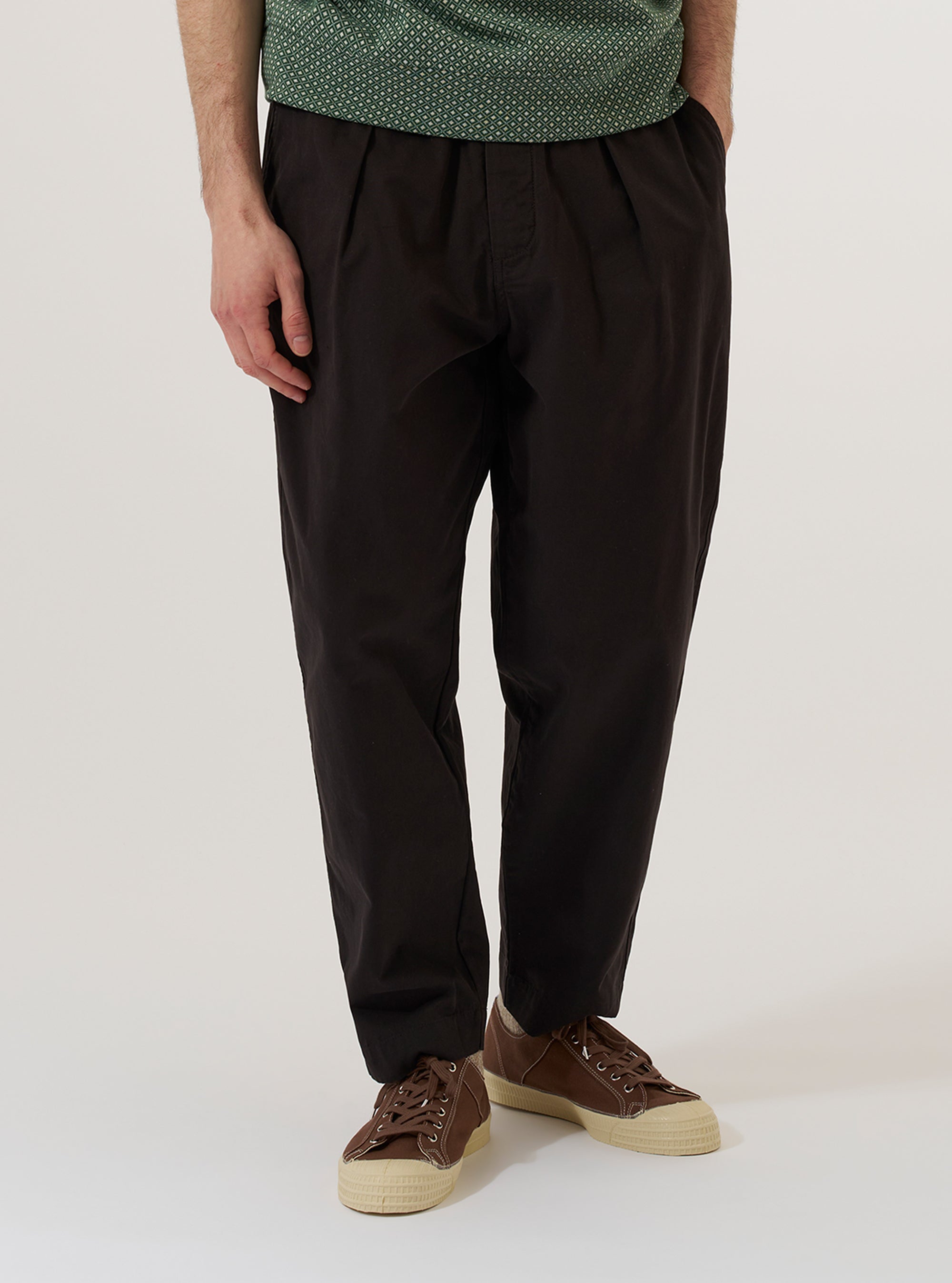 A model wears the Licorice Lyo Linen Mix Oxford Pant, shown from the waist down against a plain, neutral studio background. These loose-fit trousers in a deep brown licorice color feature a pleated front, a drawstring at the elasticated waistband, and side pockets. The model is wearing the pants with a tucked-in shirt and dark shoes, highlighting the relaxed, wide-leg silhouette.