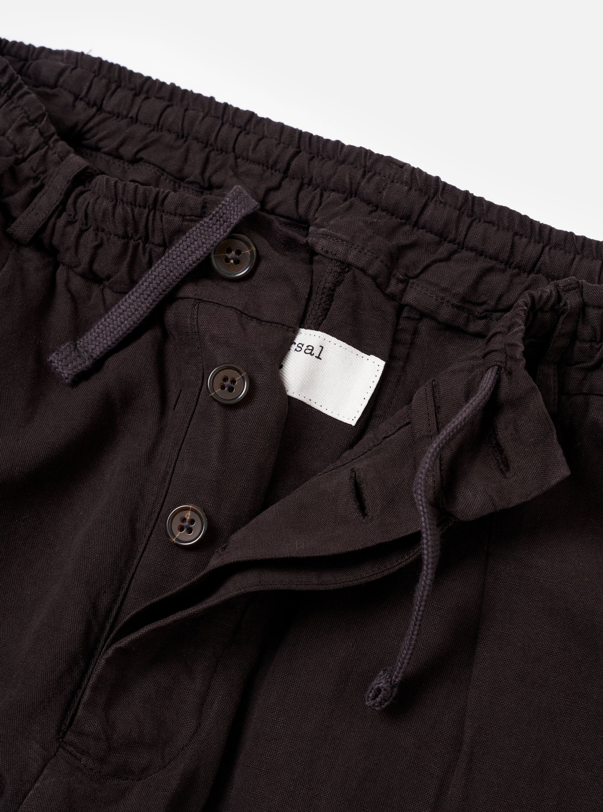 The Licorice Lyo Linen Mix Oxford Pant is shown from the back as a flat lay against a plain white background. The trousers feature an elasticated waistband and two rear pockets, including one with a buttoned flap and one button-jetted pocket. The dark brown fabric has a soft, matte appearance.