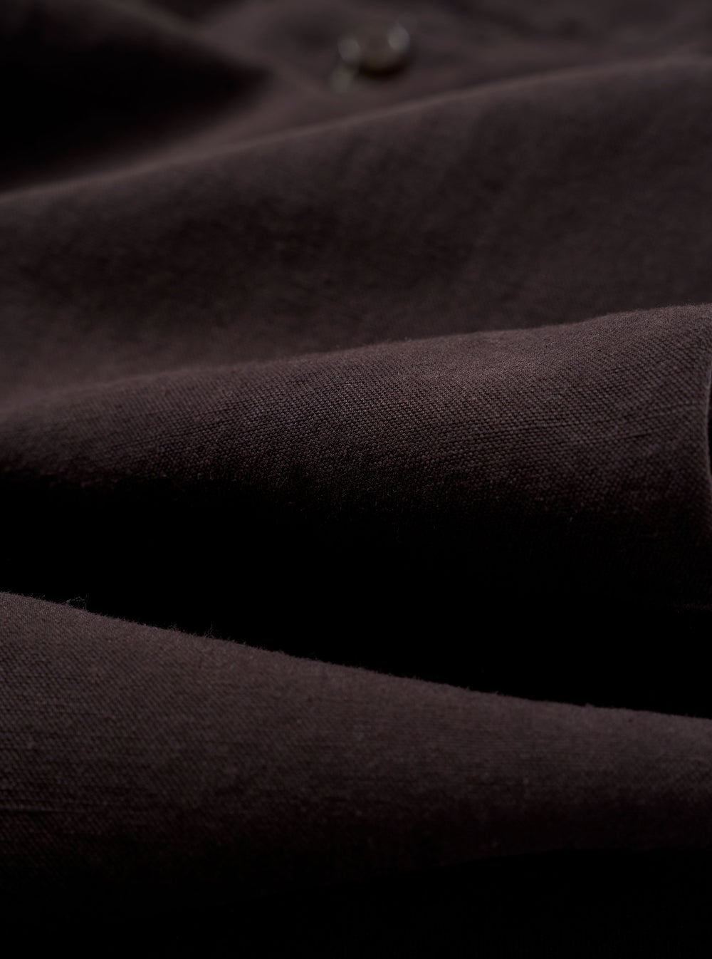 A close-up fabric swatch of the Licorice Lyo Linen Mix Oxford Pant highlights the deep brown licorice color and the subtle, natural texture of the linen and lyocell blend fabric. The image provides a detailed view of the garment's weave and material.