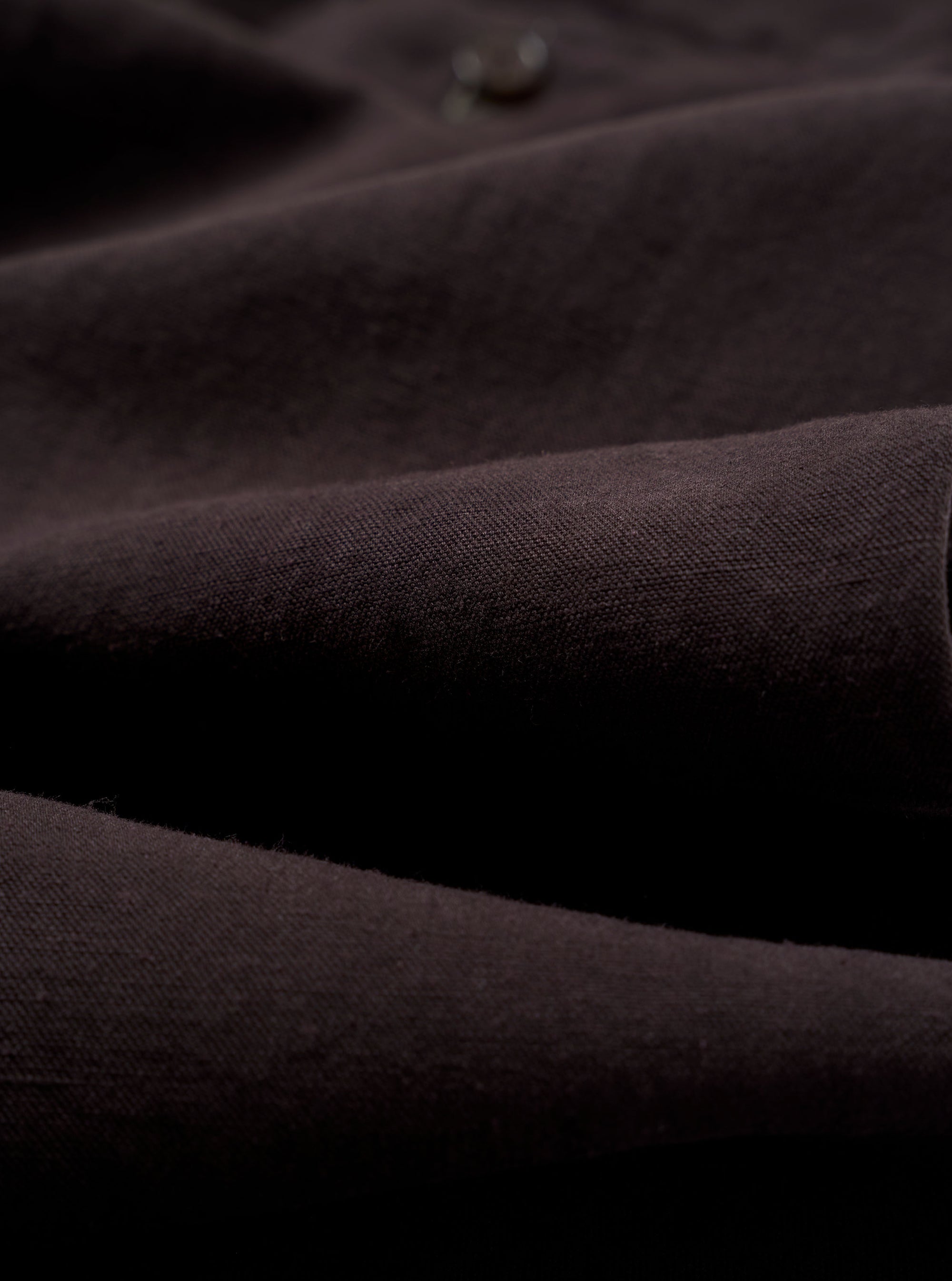 A close-up fabric swatch of the Licorice Lyo Linen Mix Oxford Pant highlights the deep brown licorice color and the subtle, natural texture of the linen and lyocell blend fabric. The image provides a detailed view of the garment's weave and material.