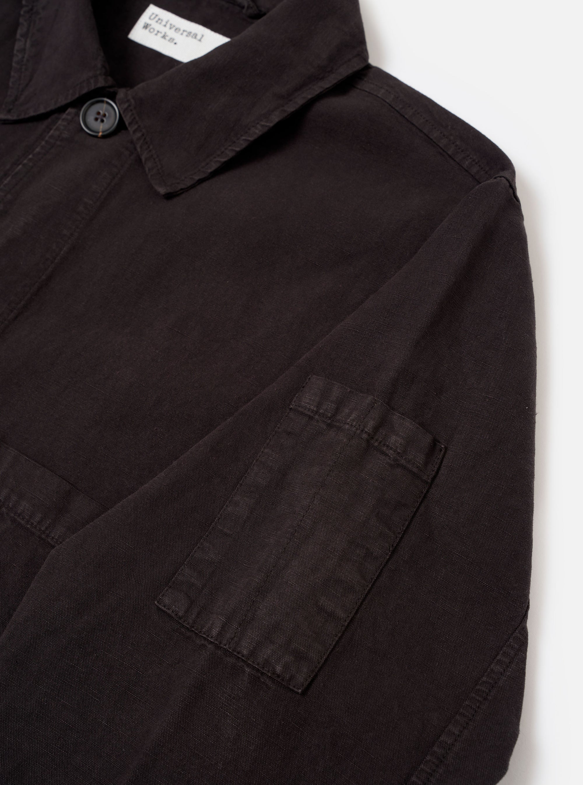 The Licorice Lyo Linen Mix Coverall Jacket is shown from the back as a flat lay against a plain white background. The image highlights the jacket’s short, square silhouette, straight hem, and simple, unbuttoned cuffs. The dark brown fabric has a visible linen-blend texture with clean stitching along the shoulders and sleeves.