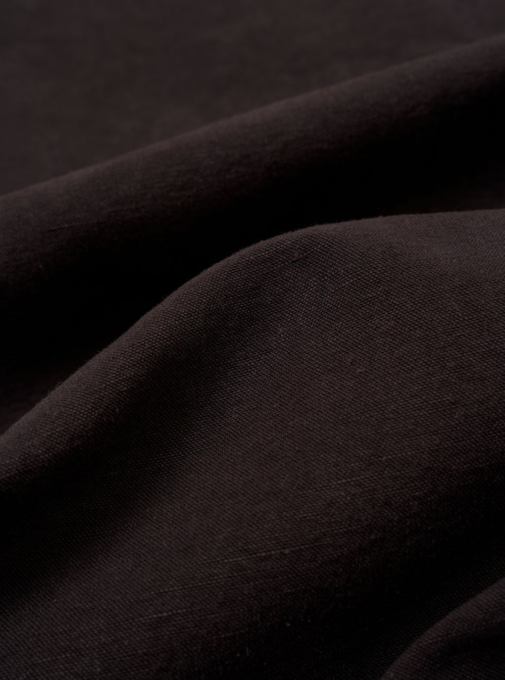 A close-up fabric swatch of the Licorice Lyo Linen Mix Coverall Jacket displays the dark brown color and the distinct texture of the linen, lyocell, and organic cotton blend. The material fills the frame, providing a detailed view of the fabric's weave.