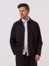 A model wears the Licorice Lyo Linen Mix Coverall Jacket against a plain, neutral background. The jacket features a classic chore-style silhouette with a structured collar, a five-button front closure, and three prominent patch pockets on the front. The fabric is a deep licorice brown lyo-linen mix with a smooth, lightweight appearance. The model is captured in a front-facing medium shot.