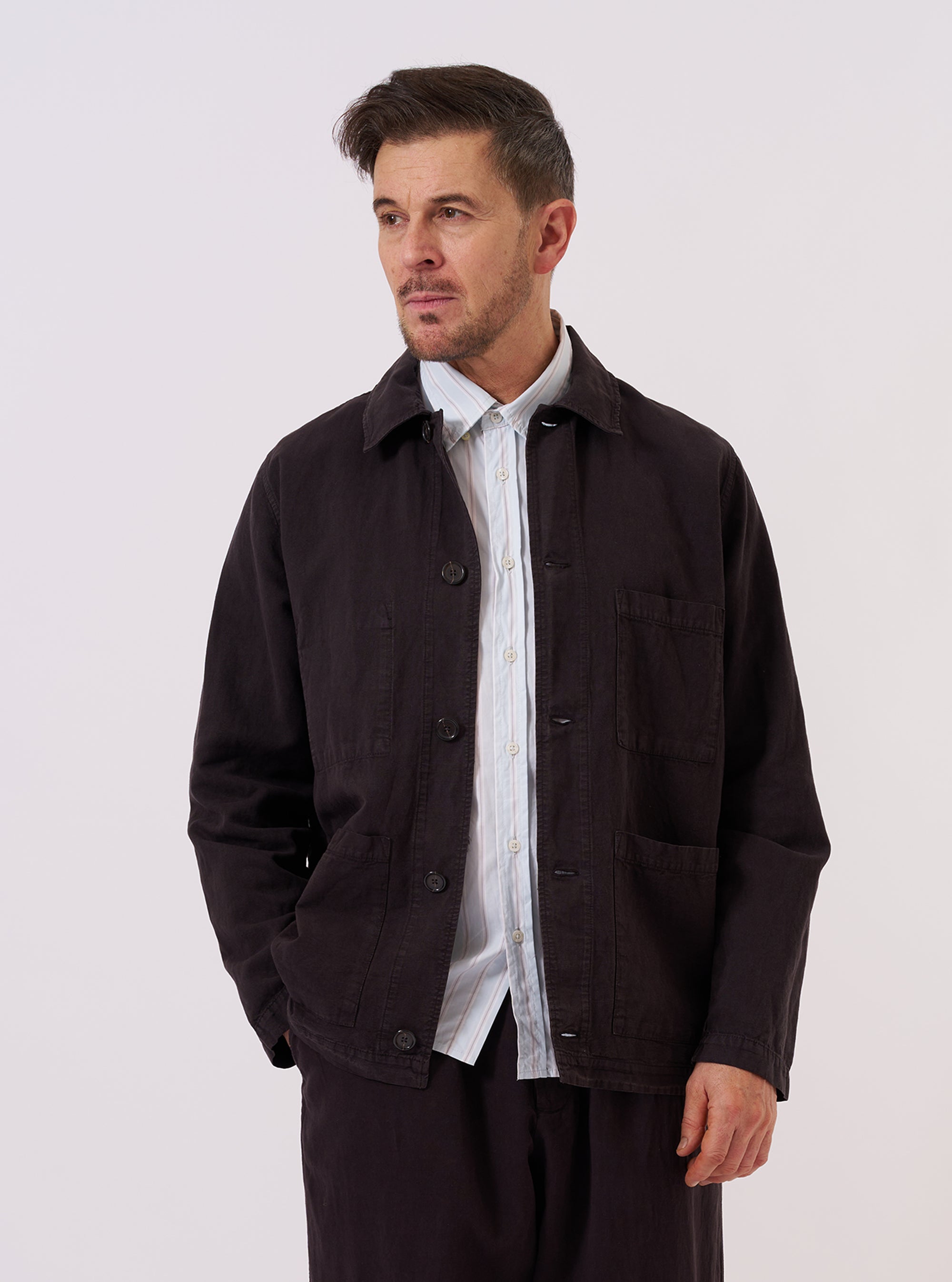 A model wears the Licorice Lyo Linen Mix Coverall Jacket against a plain, neutral background. The jacket features a classic chore-style silhouette with a structured collar, a five-button front closure, and three prominent patch pockets on the front. The fabric is a deep licorice brown lyo-linen mix with a smooth, lightweight appearance. The model is captured in a front-facing medium shot.