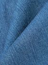 A close-up fabric swatch of the Washed Indigo 8oz Slub Denim Oxford Pant highlights the textured, irregular surface of the slub denim. The fabric is a faded indigo blue with visible white cotton fibers throughout the weave.