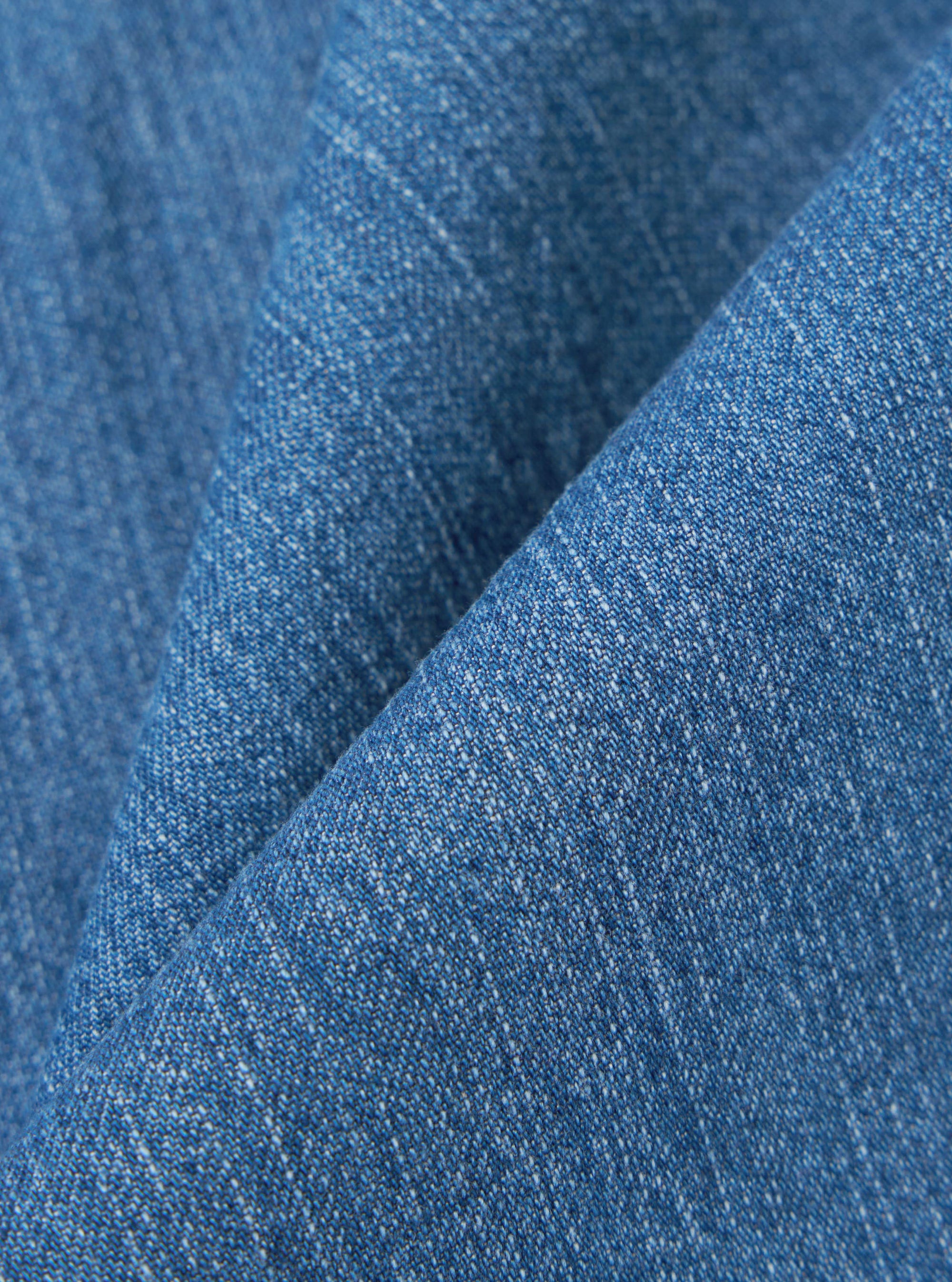 A close-up fabric swatch of the Washed Indigo 8oz Slub Denim Oxford Pant highlights the textured, irregular surface of the slub denim. The fabric is a faded indigo blue with visible white cotton fibers throughout the weave.