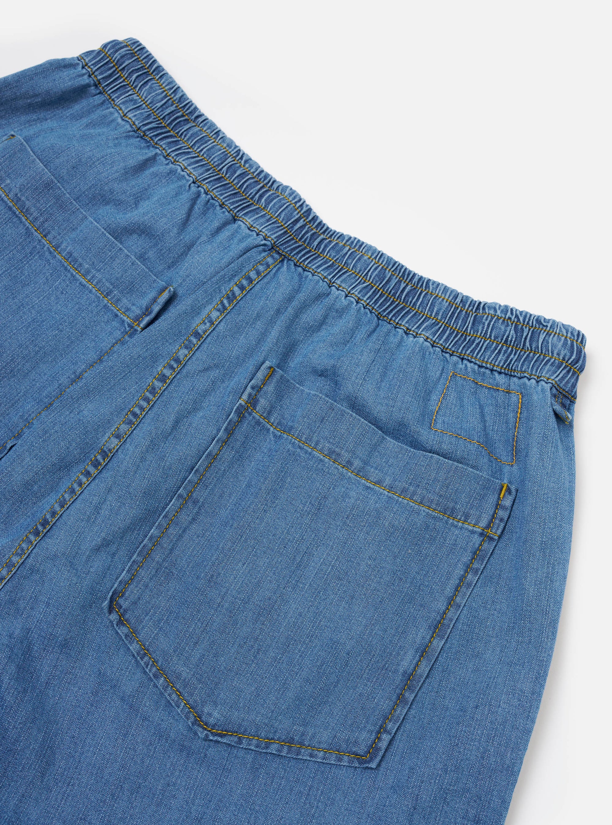 The Washed Indigo 8oz Slub Denim Hi Water Trouser is displayed from the back as a flat lay against a plain white background. The image highlights the two rear patch pockets and the elasticated waistband with a visible drawstring. The washed indigo denim fabric features a subtle slub texture and a vintage appearance.